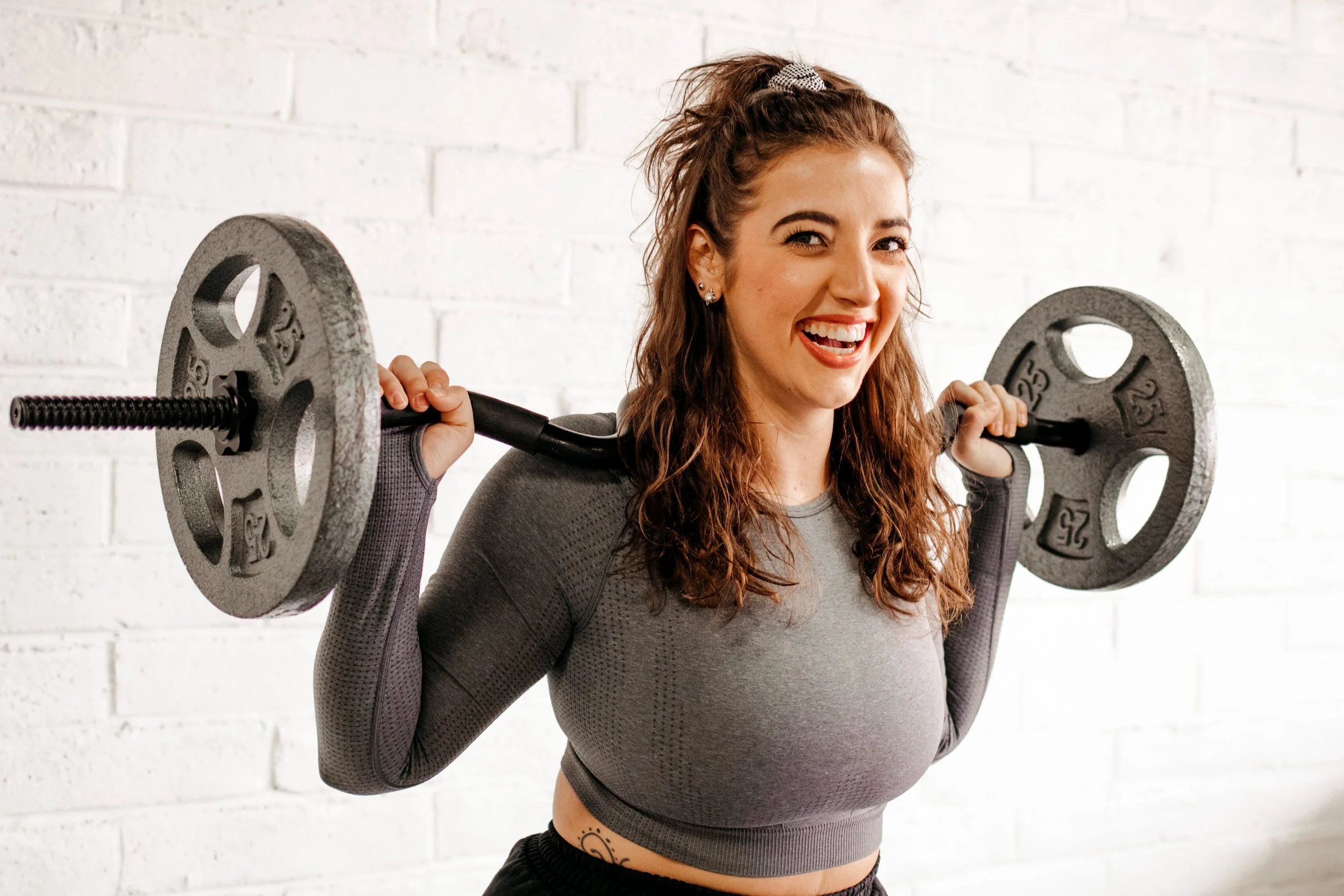 A young woman with curly red hair in a high ponytail, wearing a gray athletic long sleeve shirt, is lifting a barbell with weight plates on each end. She is smiling and standing in front of a white brick wall.