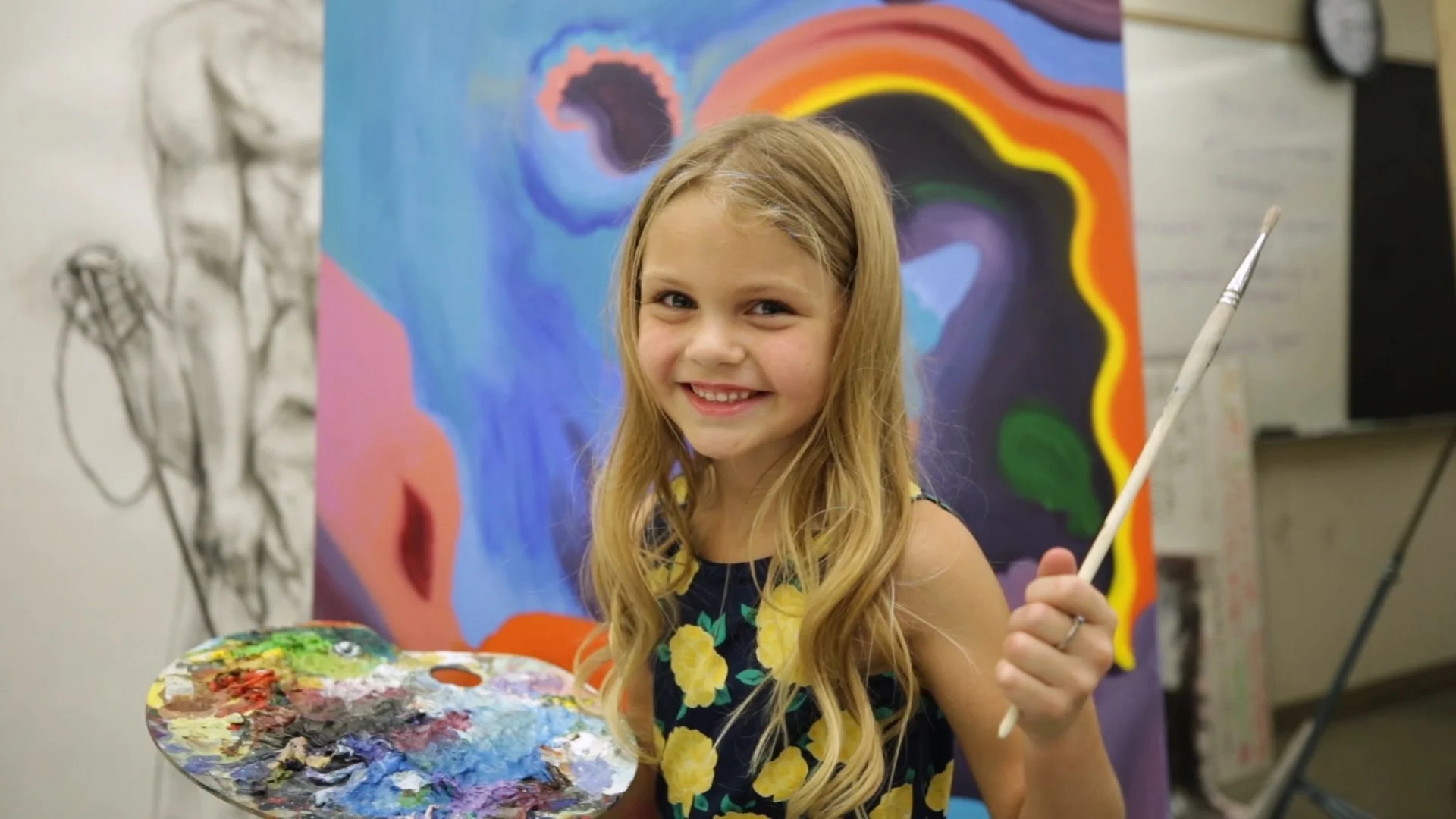 A young girl with long blonde hair smiling and holding a paintbrush in front of a colorful abstract painting she is creating in an art classroom.