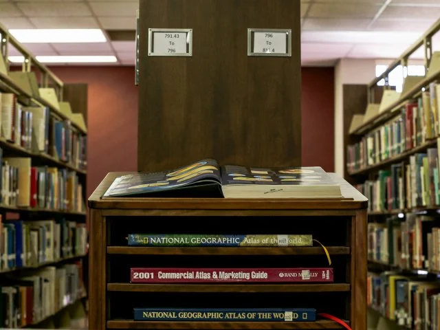 A library with bookshelves, a wooden table with open books on it, and labeled aisle signs overhead.
