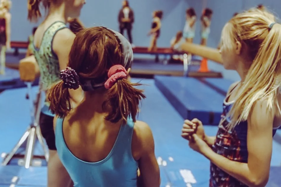 Two young girls talking in a gymnastics training facility, with other gymnasts practicing on balance beams in the background.