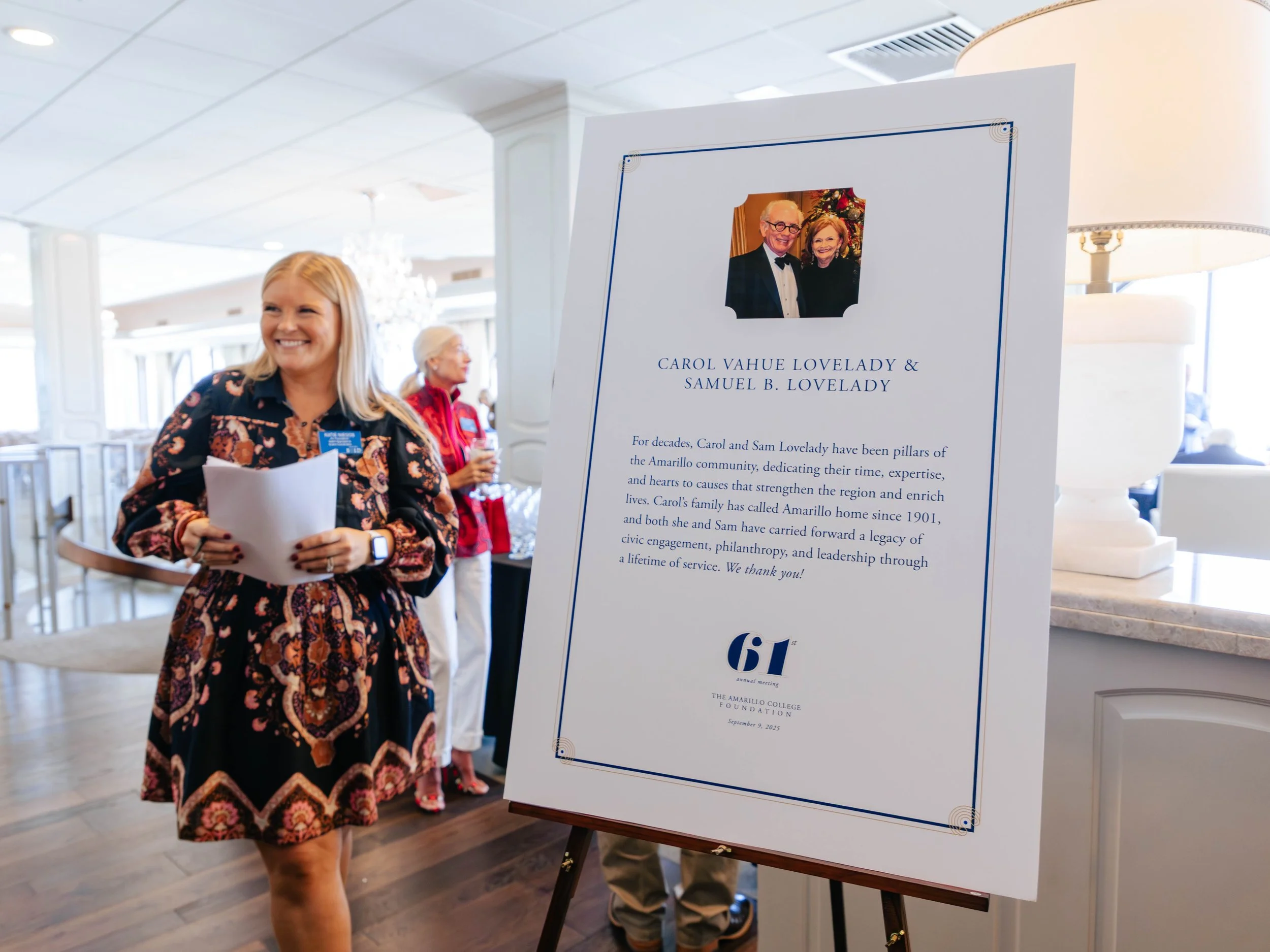 A woman in a patterned dress smiling while holding papers next to a sign contains text about Carol Vahue Lovelady and Samuel B. Lovelady and mentions the Amarillo College Foundation.