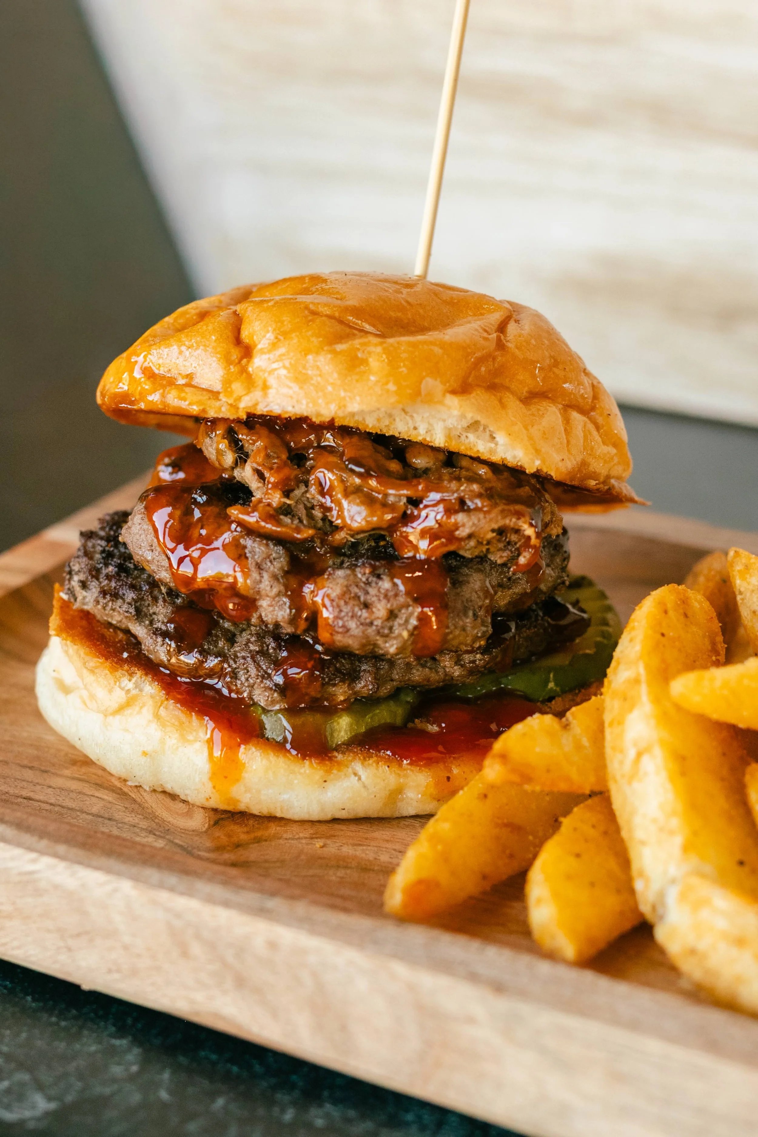 Close-up of a brisket cheeseburger with toppings and sauce, served with French fries on a wooden platter.