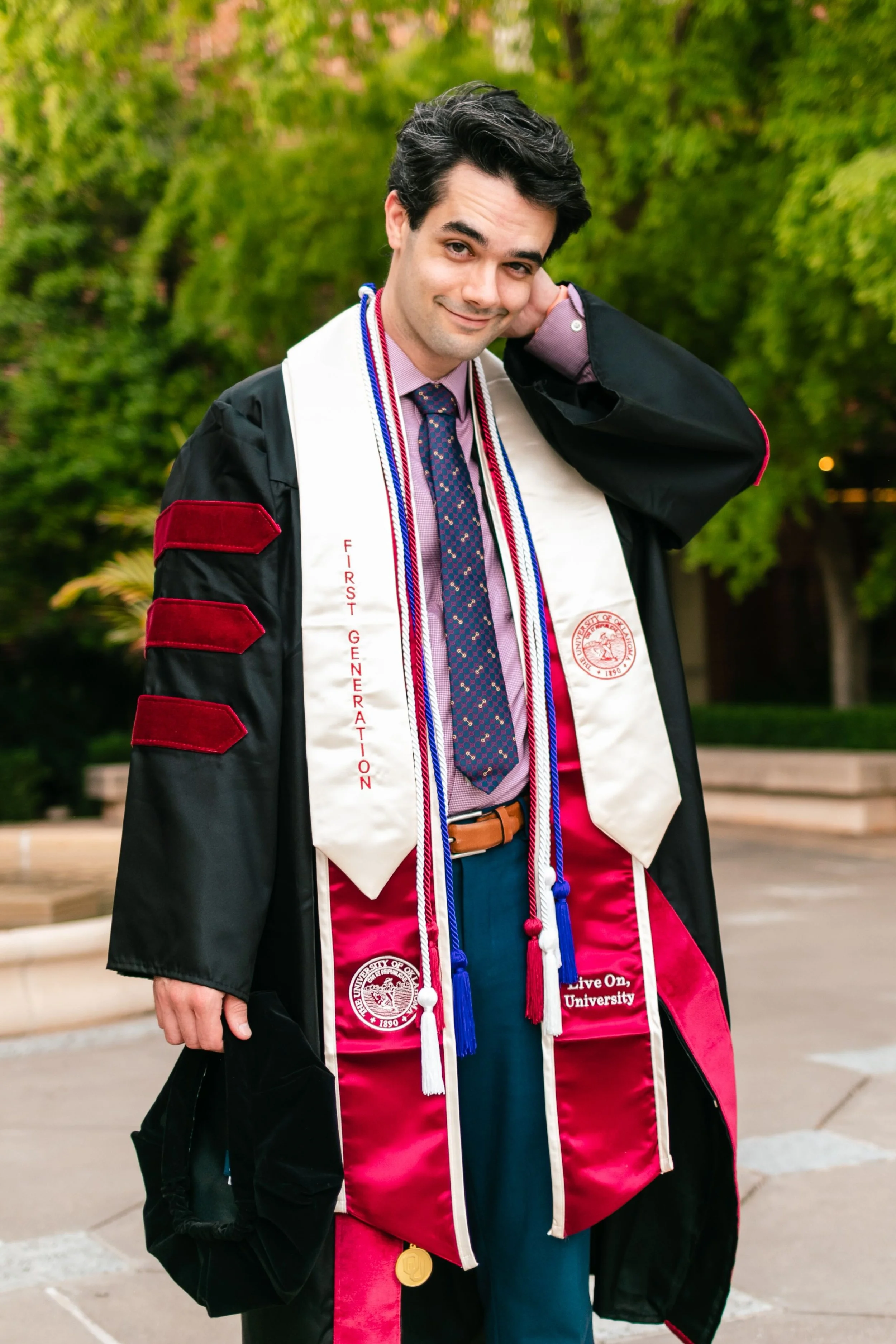 A man in his law school graduation gown and stole, outdoors with green trees in the background, smiling and posing for a graduation photo.