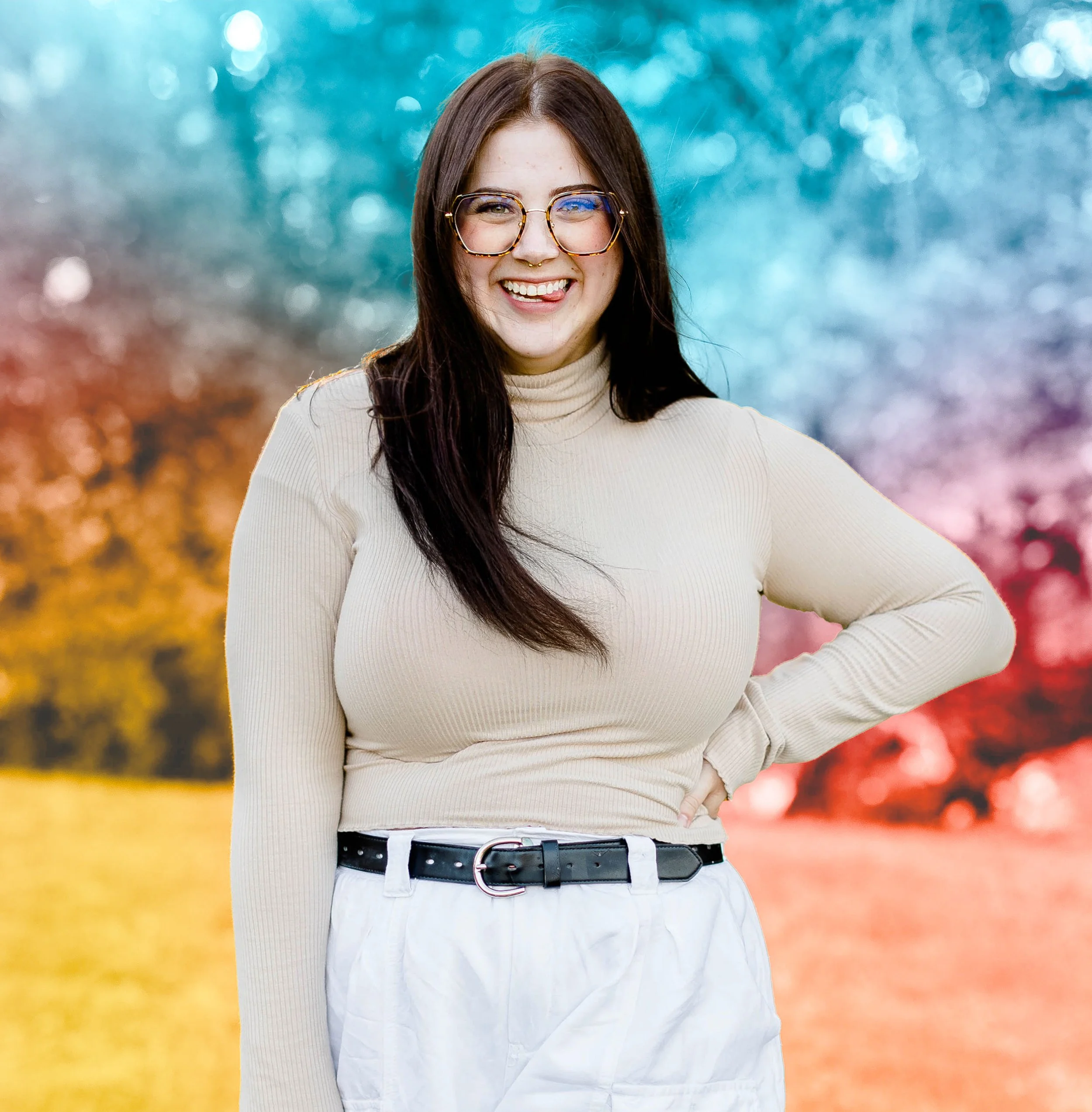 A portrait of Lily K Gamble with long dark hair, wearing glasses, a beige turtleneck sweater, and white pants with a black belt, standing outdoors with blurred colorful autumn trees in the background.