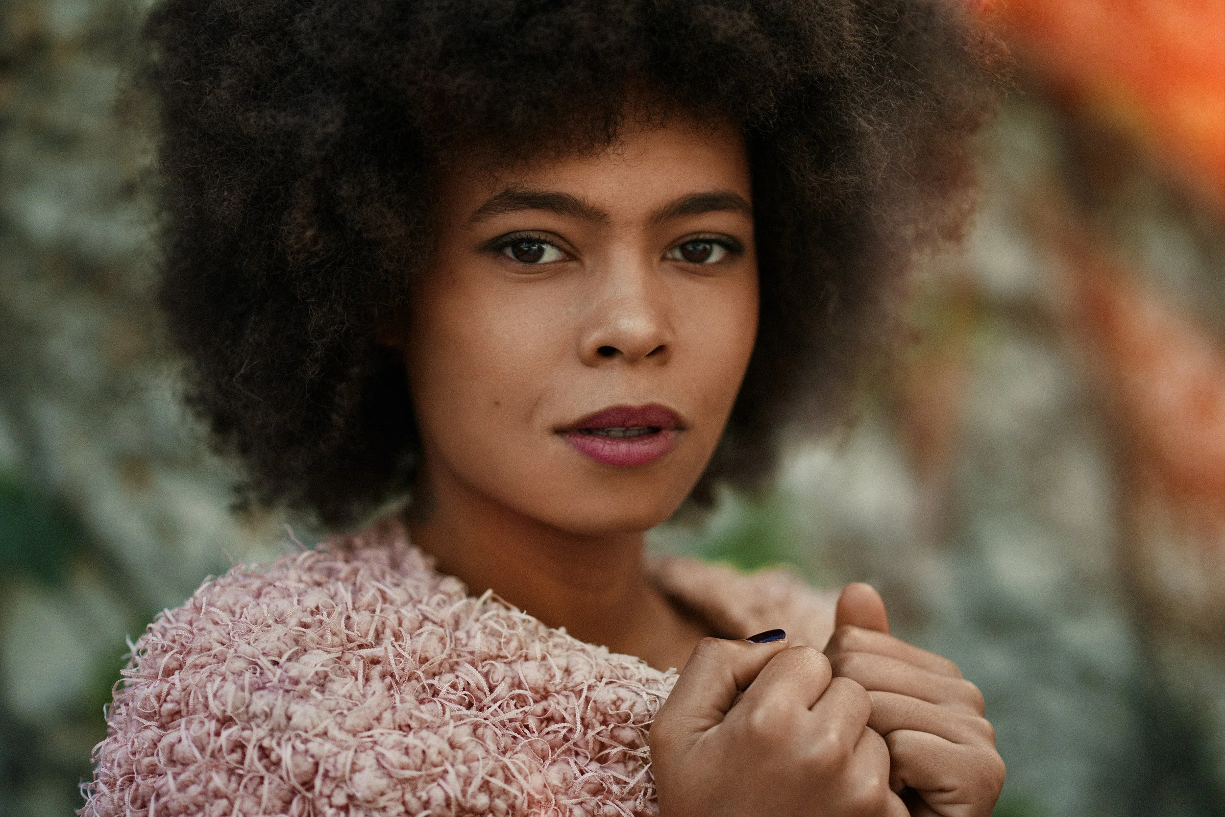 Close-up of a young woman with dark skin and an afro hairstyle, wearing a pink, textured sweater, standing outdoors with blurred greenery in the background.