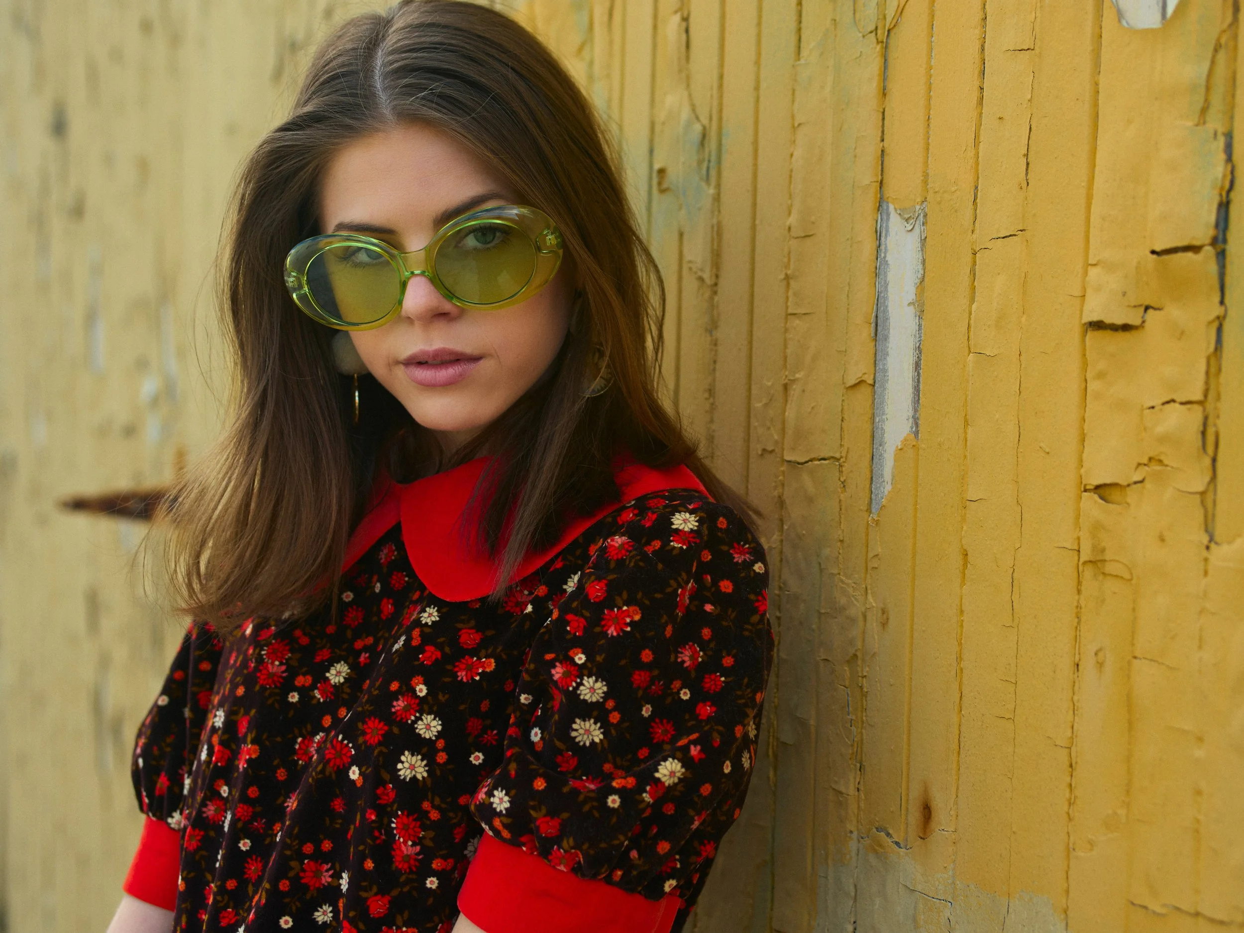 A young woman with brown hair and green eyes wearing large, reflective green sunglasses and a black dress with red and white floral pattern, leaning against a yellow brick wall.
