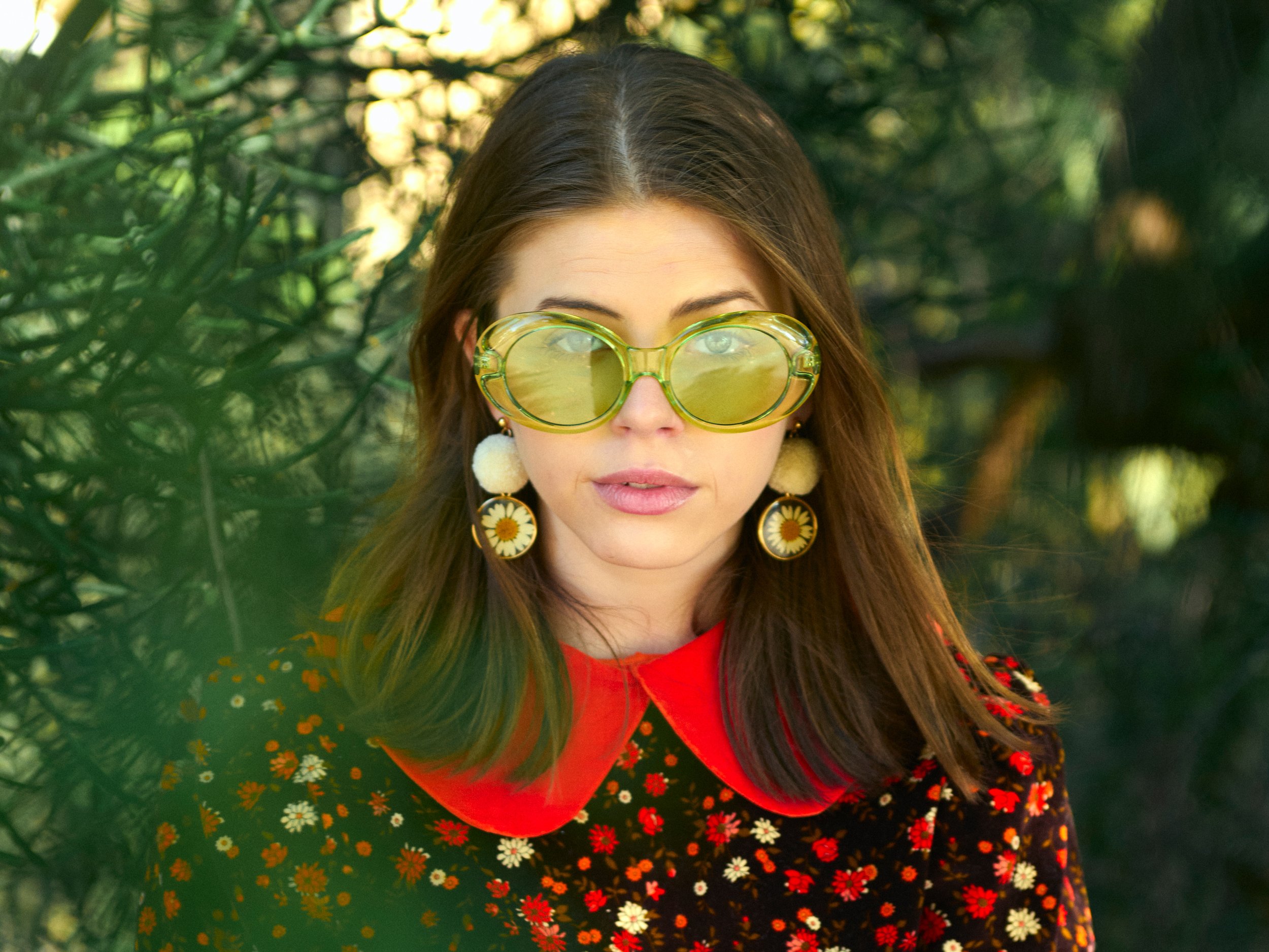 A woman with long brown hair wearing yellow retro sunglasses, large daisy flower earrings, a red top, and a floral dress with daisy designs, standing outdoors among green trees and plants.