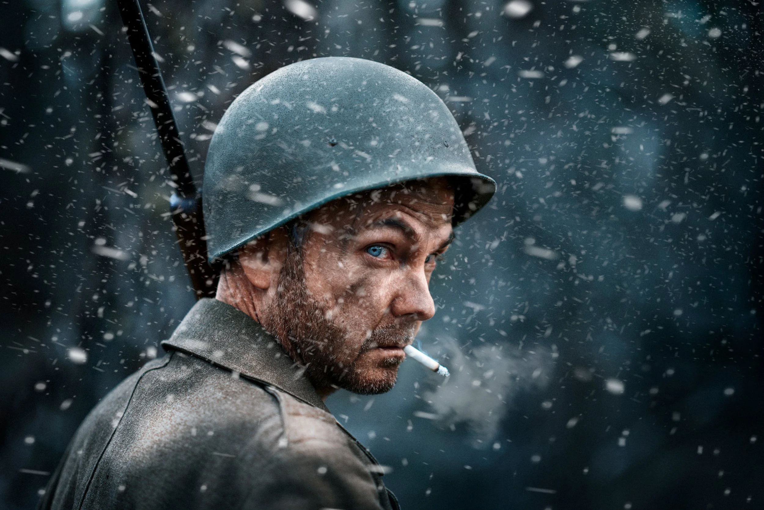 A man wearing a military helmet with a stern expression, a cigarette in his mouth, in a snowy environment with snow falling around him.