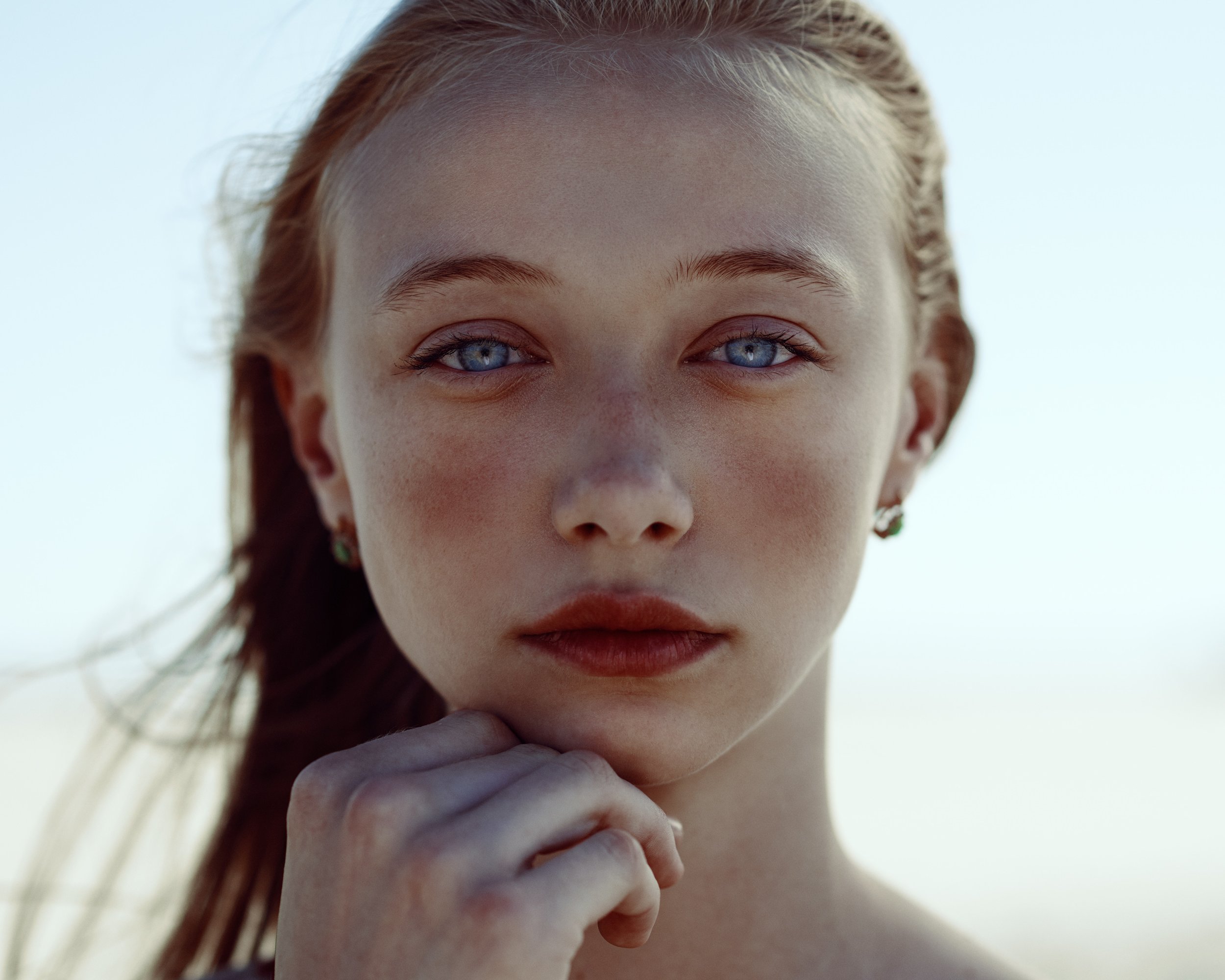 Close-up of a young woman with blue eyes, red hair, and fair skin, looking directly at the camera against a bright sky background.