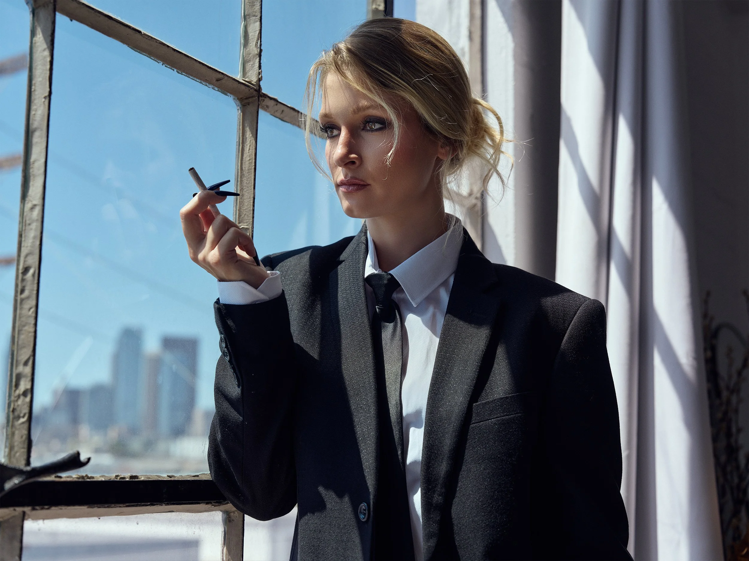 A woman in a business suit holding a cigarette and looking outside through a window with a cityscape in the background.