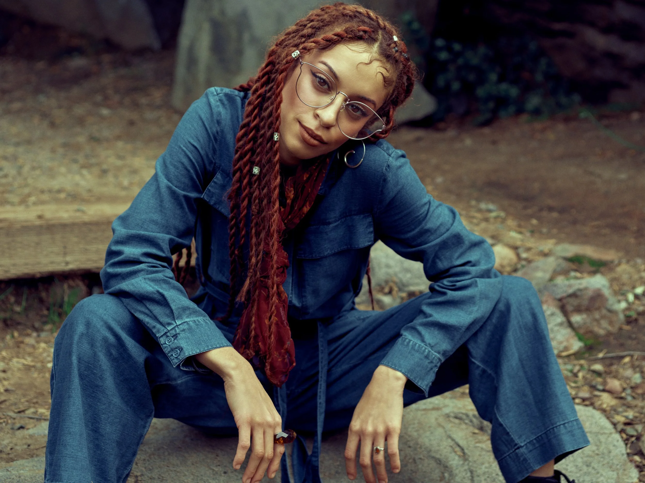 A woman with red braided hair, glasses, and jewelry wearing a denim shirt, sitting outdoors in a rocky, forested area.