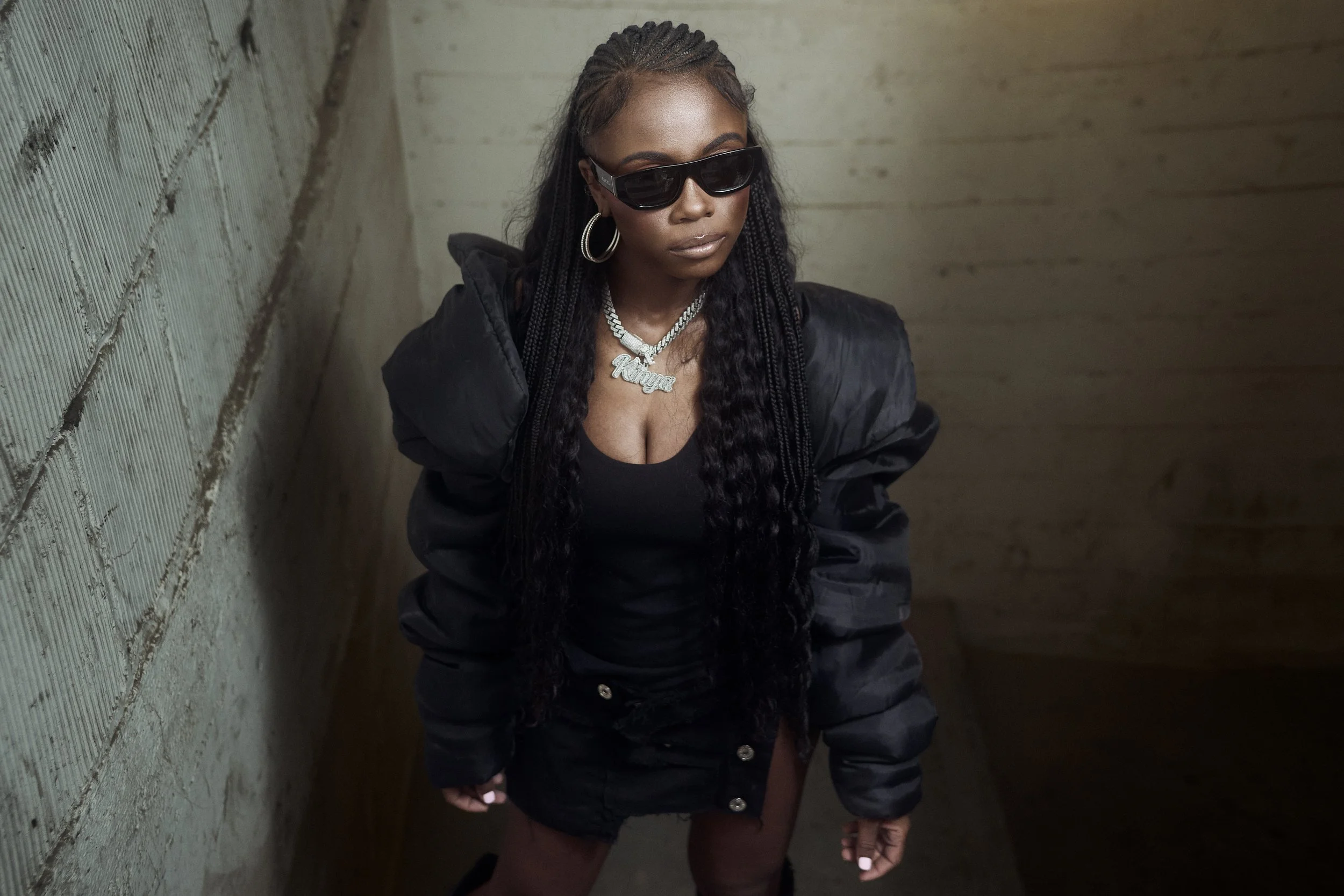 A woman with long braided hair wearing sunglasses, hoop earrings, a silver necklace, a black top, and a black jacket, standing against a concrete wall.