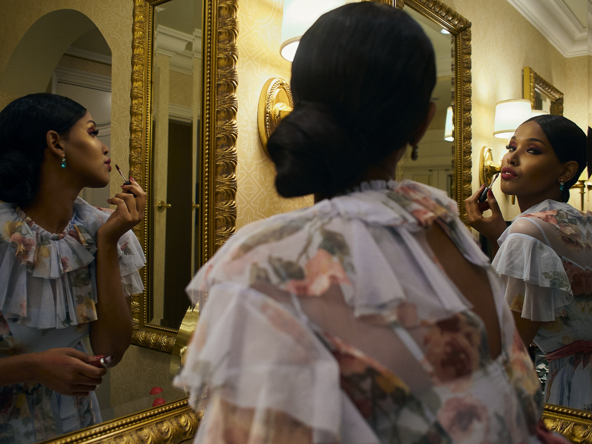 A woman with dark hair and makeup applying lipstick while looking in a large ornate gold-framed mirror. She is wearing earrings and a floral blouse. The mirror is on a yellow patterned wall with wall lamps reflected in the mirror.