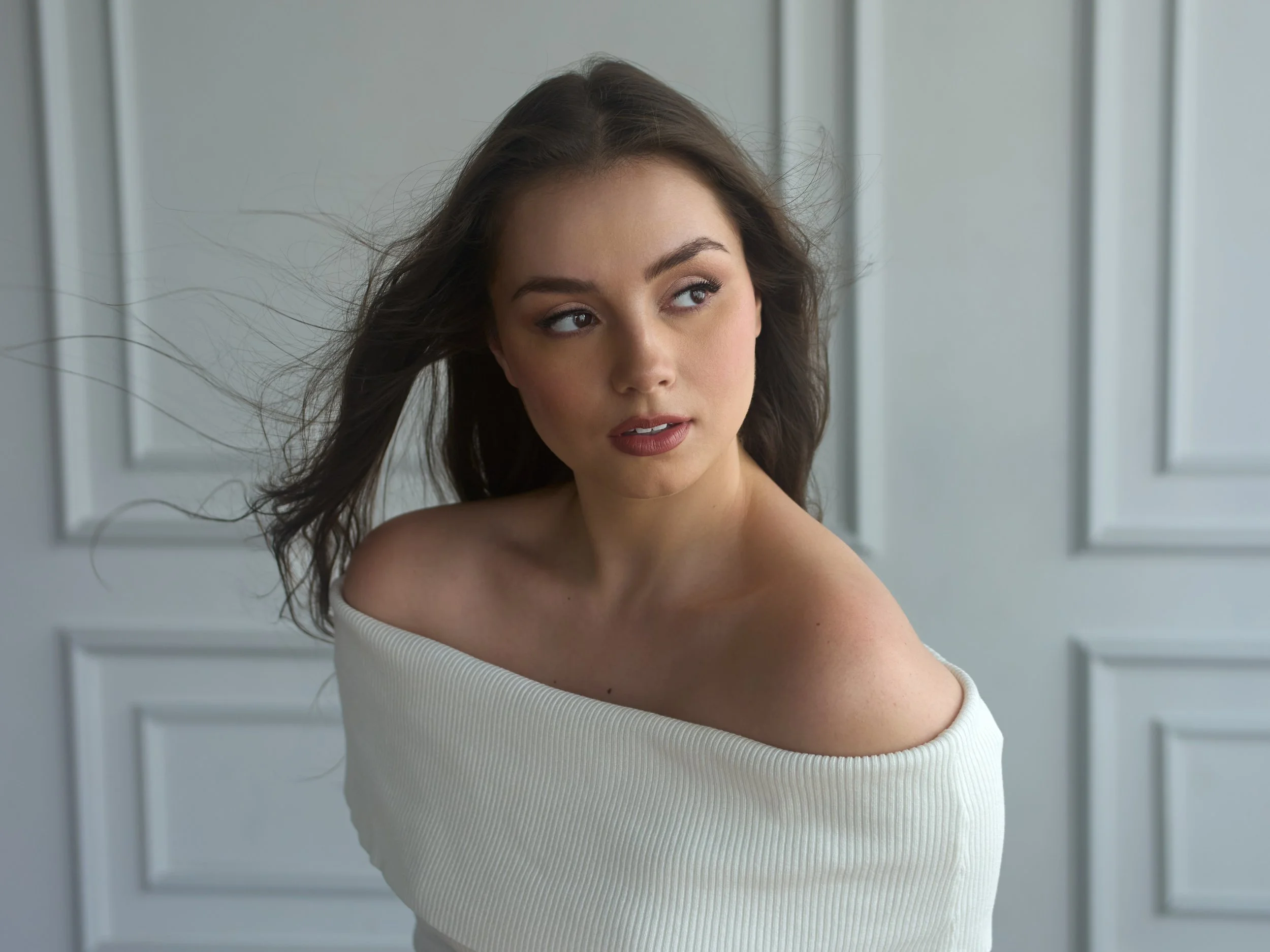 A young woman with long, wavy brown hair looking to her left. She is wearing an off-the-shoulder white ribbed top, with wind blowing her hair. The background is light gray with decorative molding.