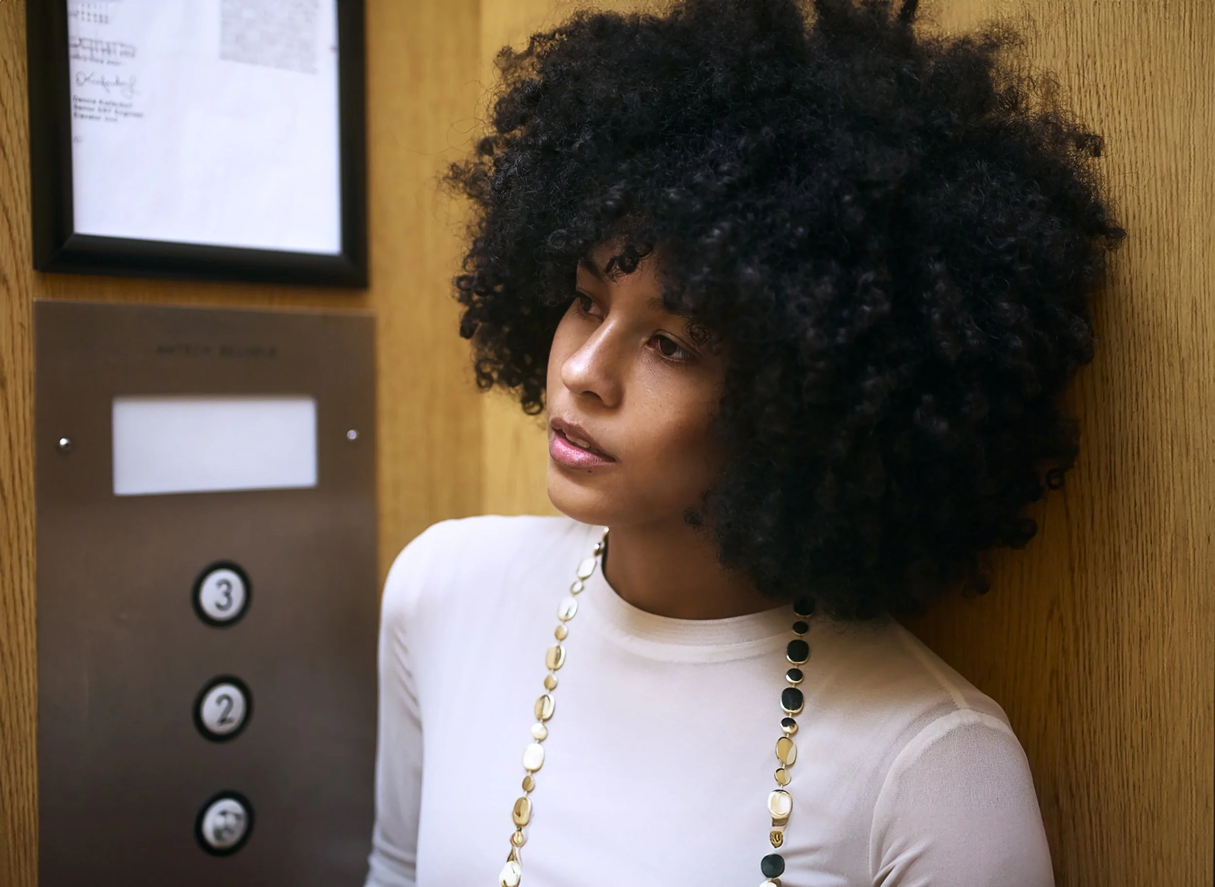 A woman with curly black hair and light skin wearing a white top and a long necklace with black and gold beads, standing beside an elevator panel with four black numbered buttons, on a wooden wall.