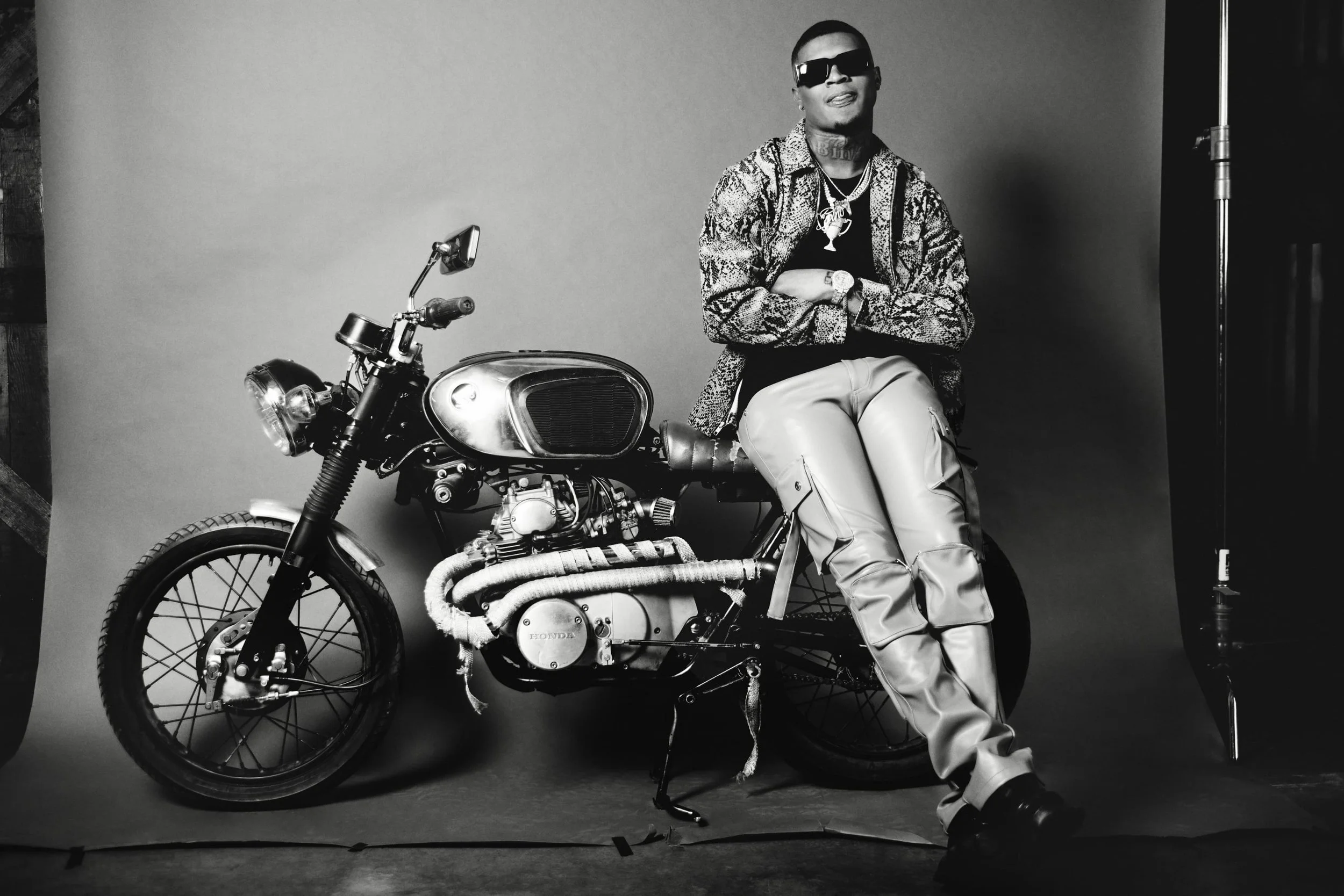 A man wearing sunglasses, a snake-patterned jacket, and light-colored pants, sitting on a vintage motorcycle in a studio setting with a plain background.