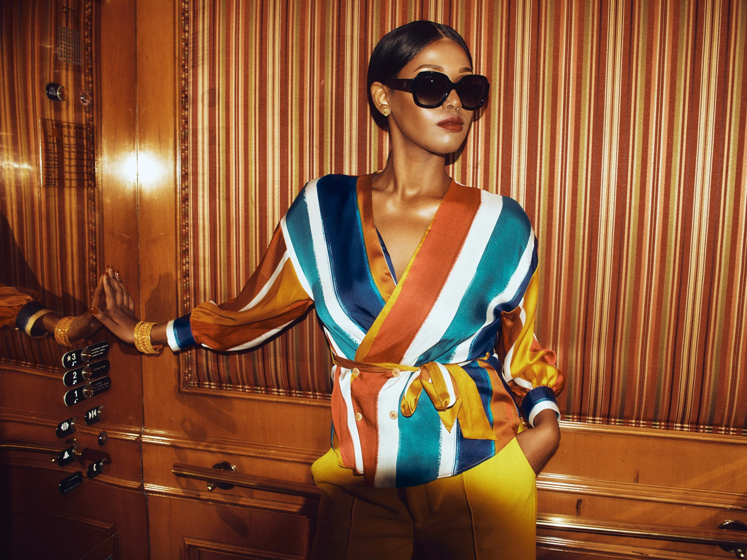 A woman with short black hair wearing oversized sunglasses, a colorful striped blouse, and yellow pants standing inside an elevator with wood paneling and a control panel visible.