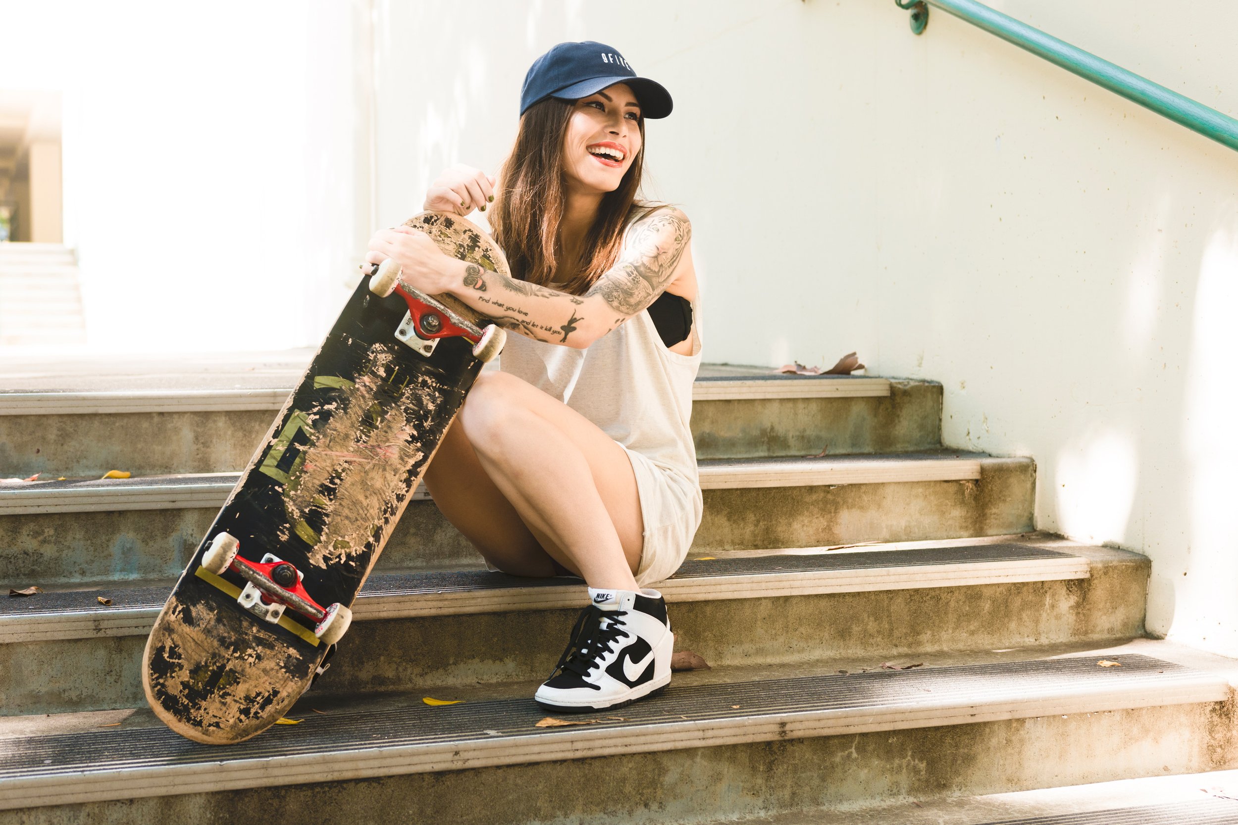 Young woman with tattoos sitting on steps, smiling, holding skateboard, wearing a black baseball cap, beige shorts, and sneakers.