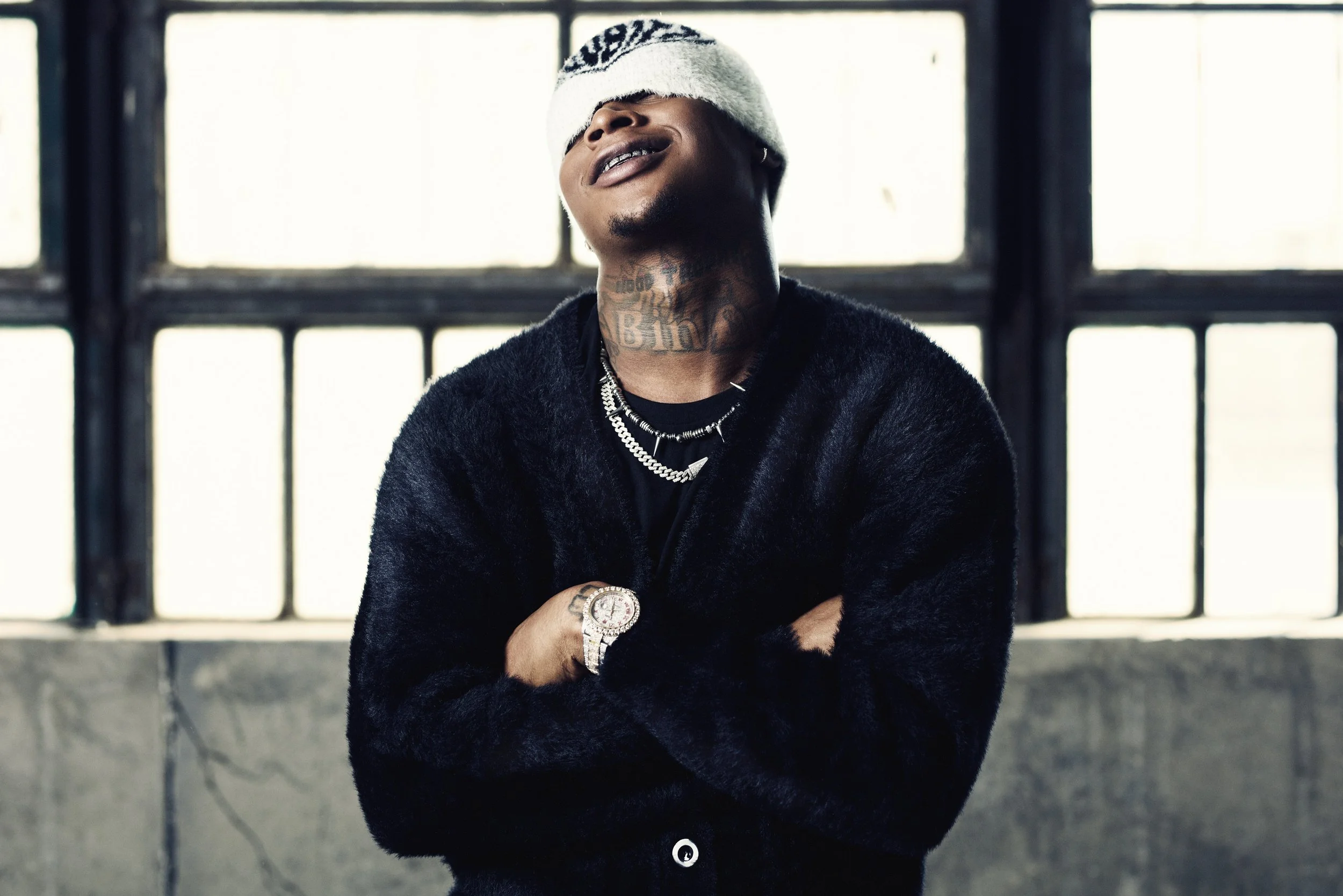 A young man with visible tattoos on his neck, wearing a white beanie and multiple chains, stands with his arms crossed in front of large windows, smiling with visible grills.