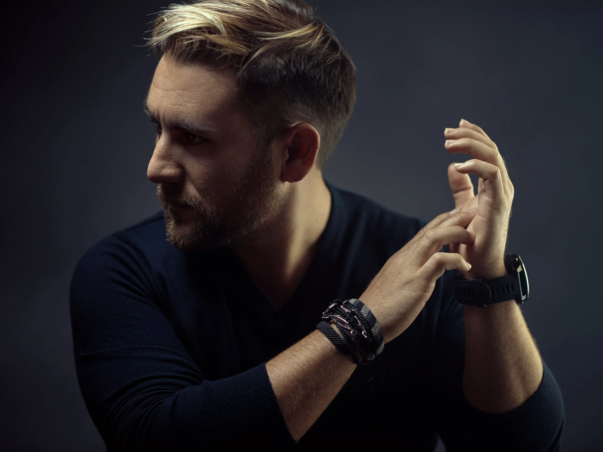 A young man with a beard and styled hair, wearing multiple bracelets, in profile against a dark background.