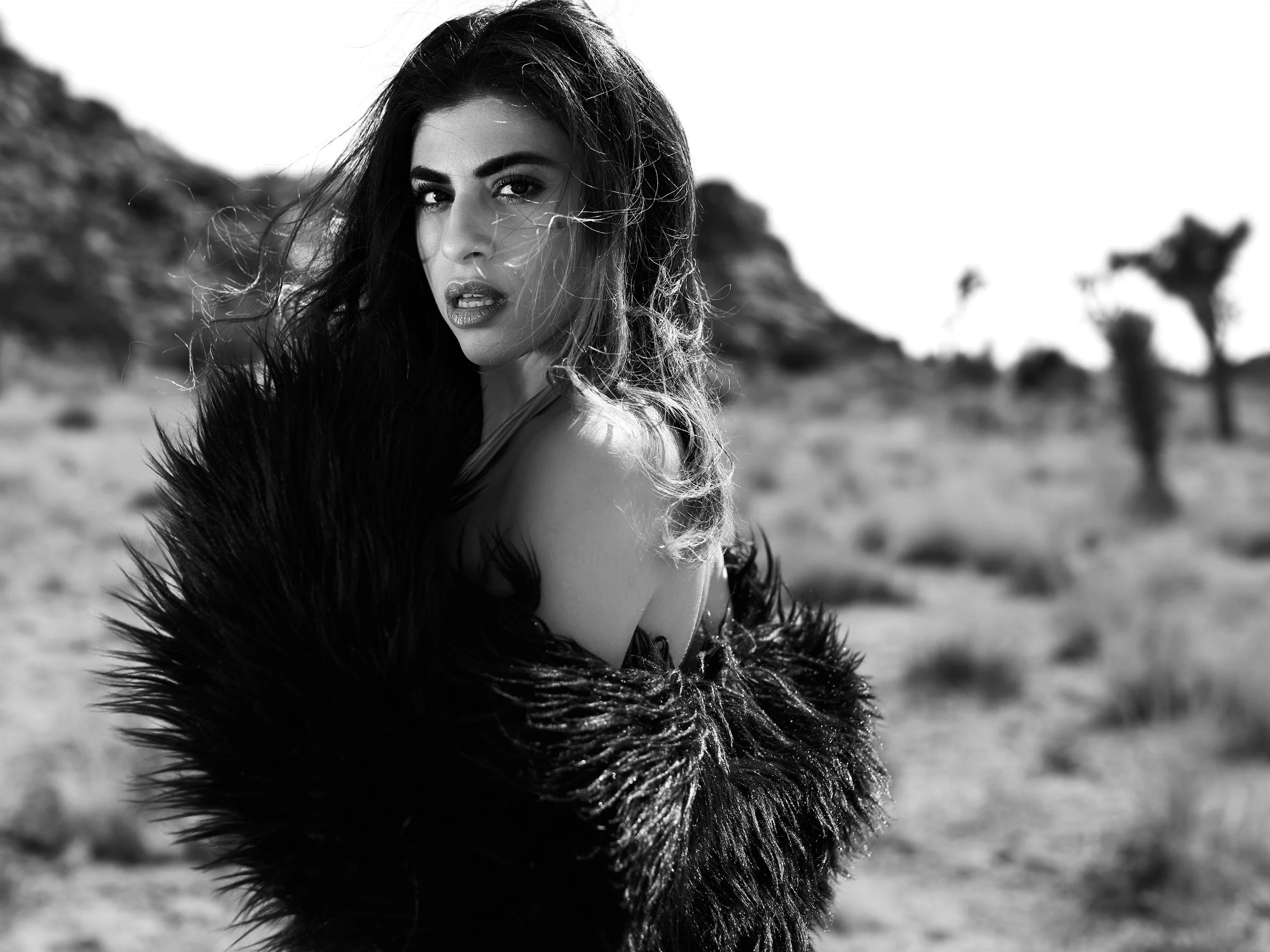 A black and white portrait of a woman with long, dark hair and a serious expression, standing outdoors with blurred trees and landscape in the background.