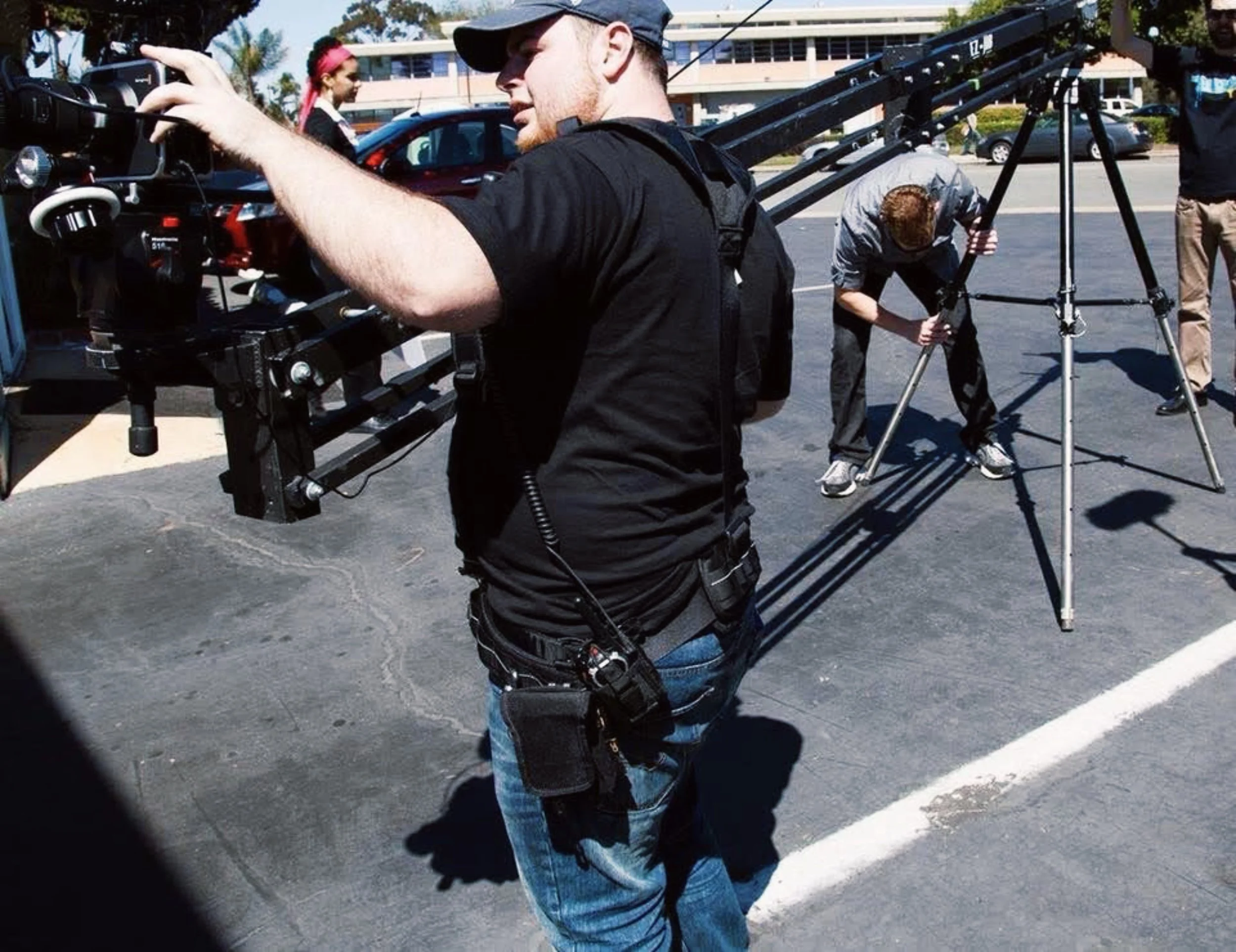 A man wearing a black shirt and jeans is operating a camera mounted on a shoulder rig outside in a parking lot, with a woman in a gray hoodie and pink headband in the background, and a person working on camera equipment nearby.