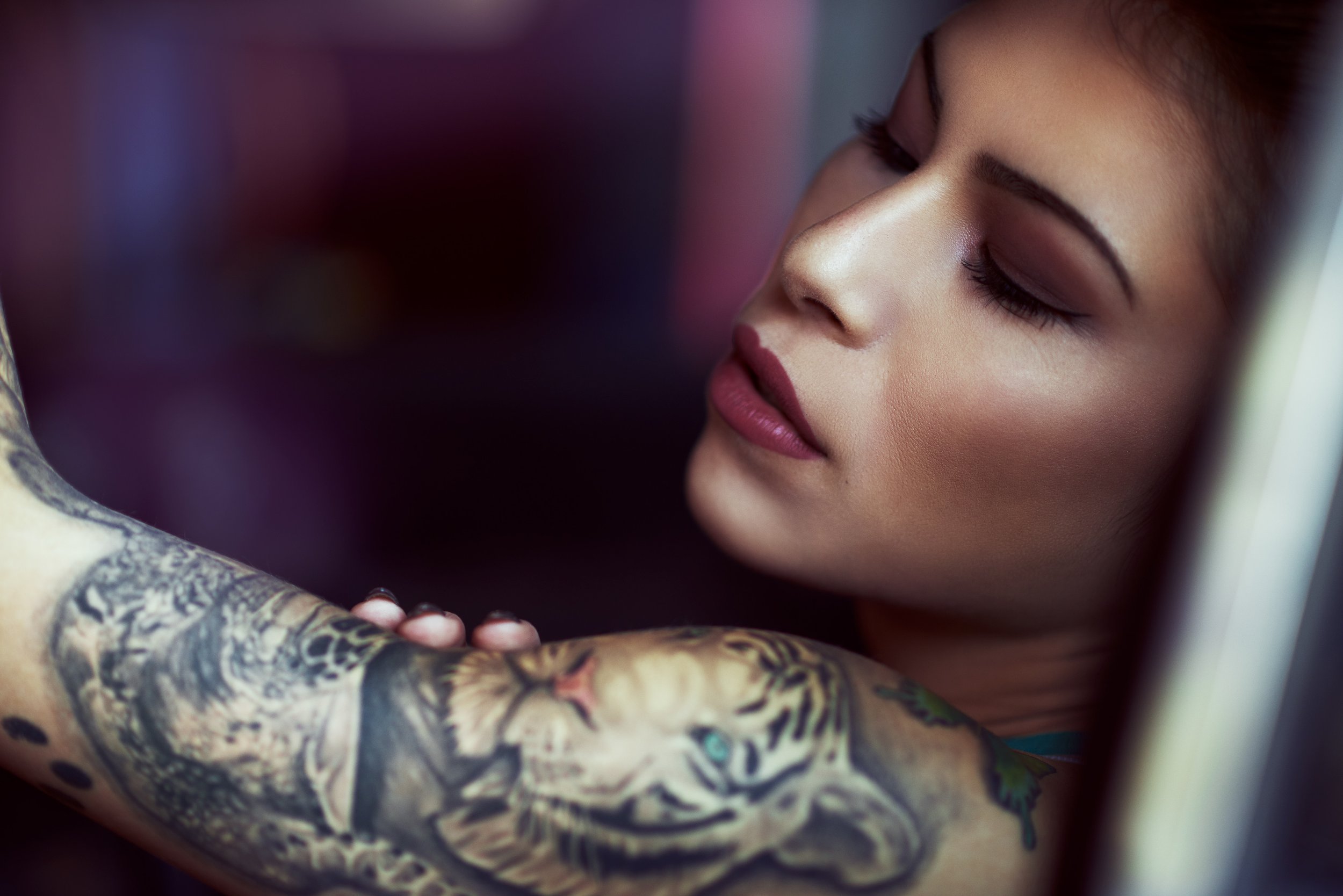 A woman with makeup and tattoos on her arm, her eyes closed, leaning against a wall or surface.