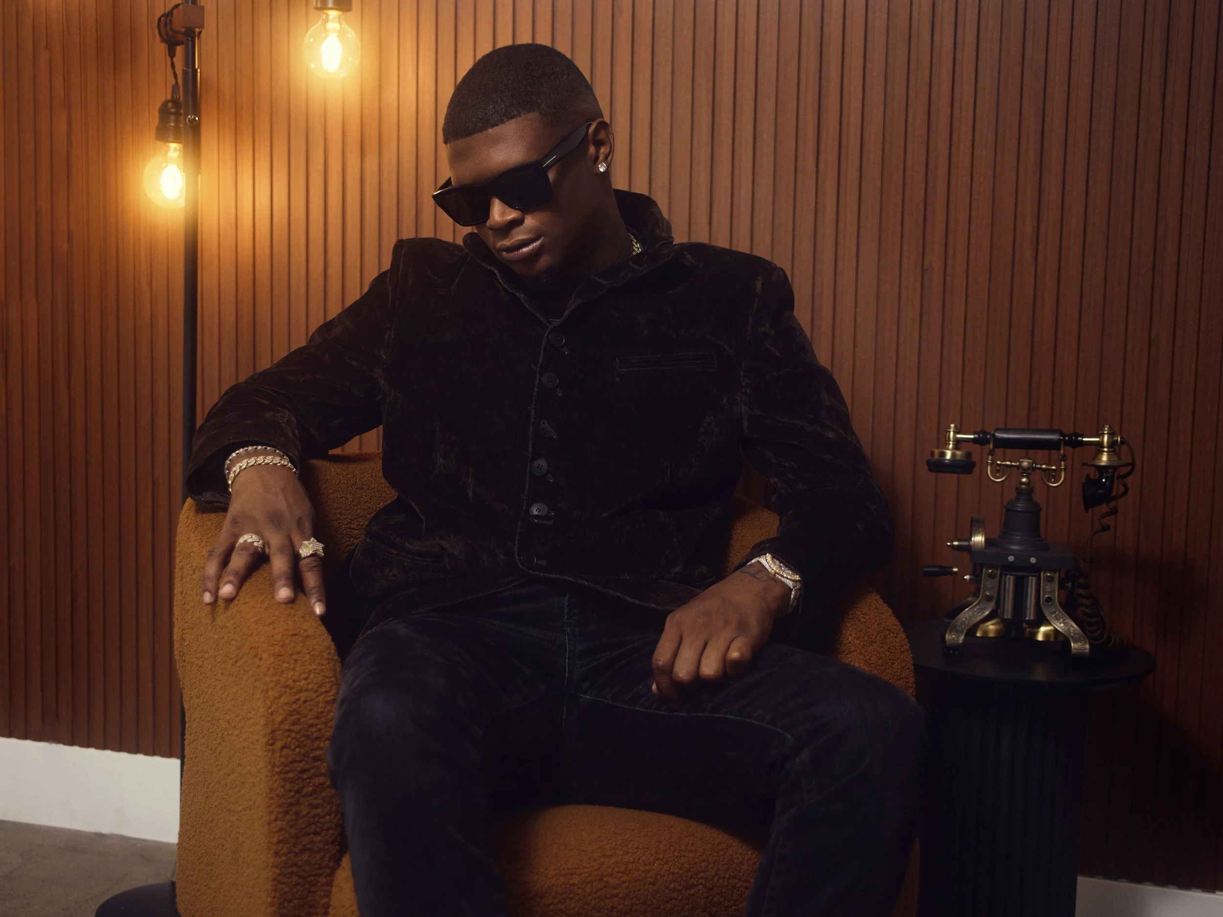 A young man with dark skin wearing sunglasses and jewelry sitting on an orange upholstered chair in front of a wooden-paneled wall, with two exposed light bulbs hanging on the left and a vintage microscope on the right side of the image.