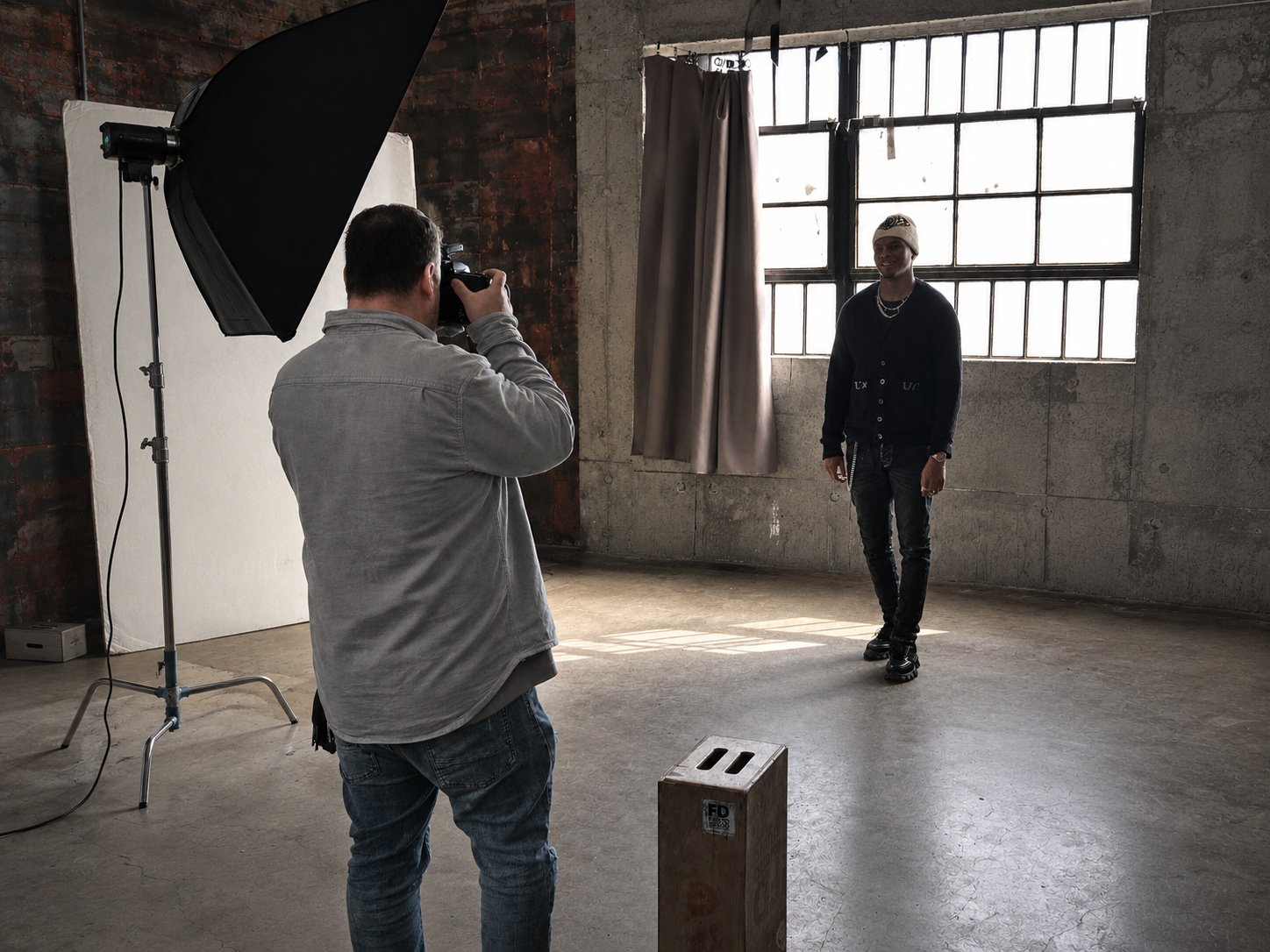 A photographer takes a picture of a young man posing in an industrial-style studio with concrete walls and large windows.