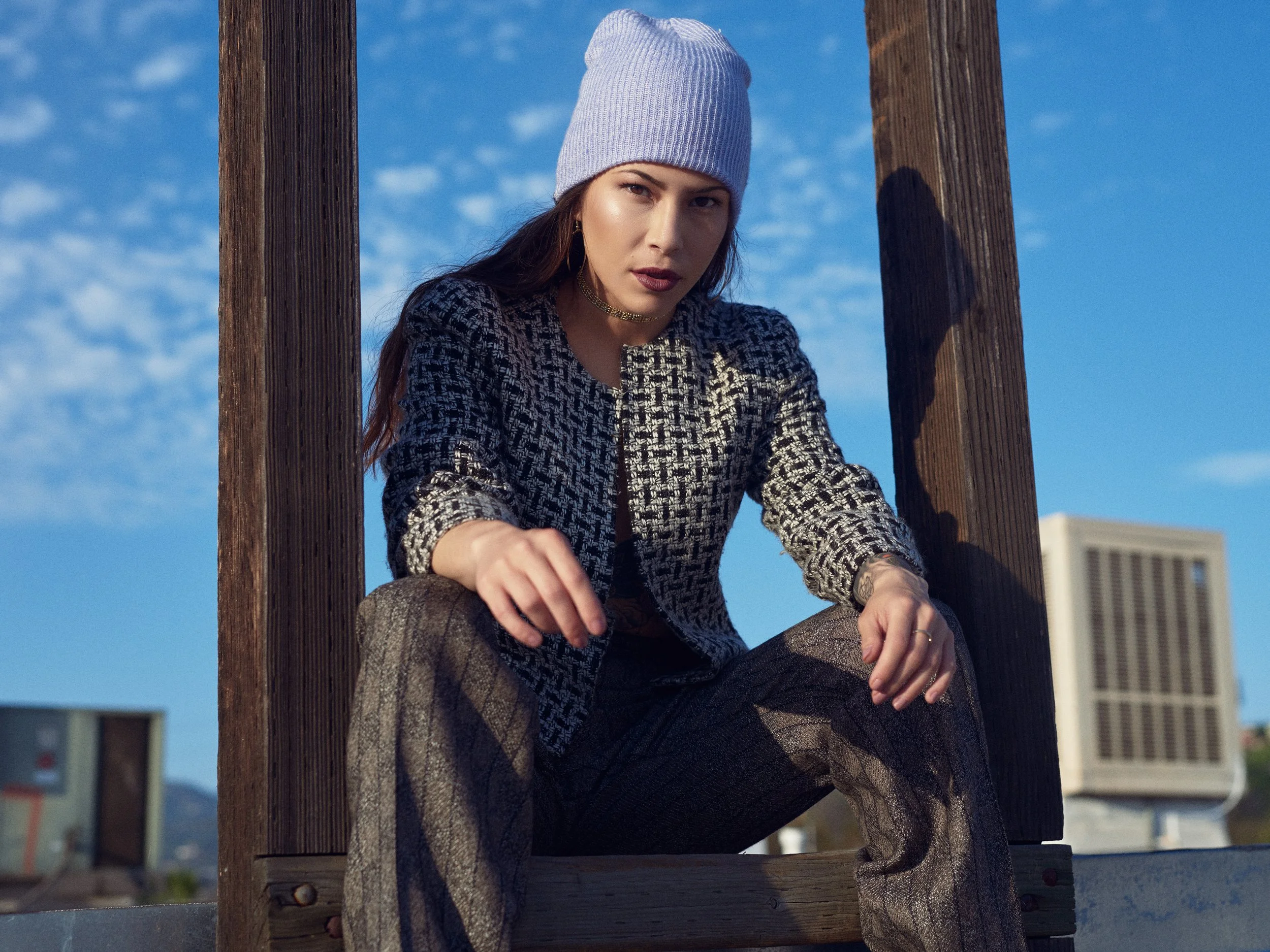 A woman with long brown hair wearing a light gray beanie, a black and white patterned jacket, and gray pants, sitting outdoors between two wooden beams, with a cityscape and blue sky in the background.