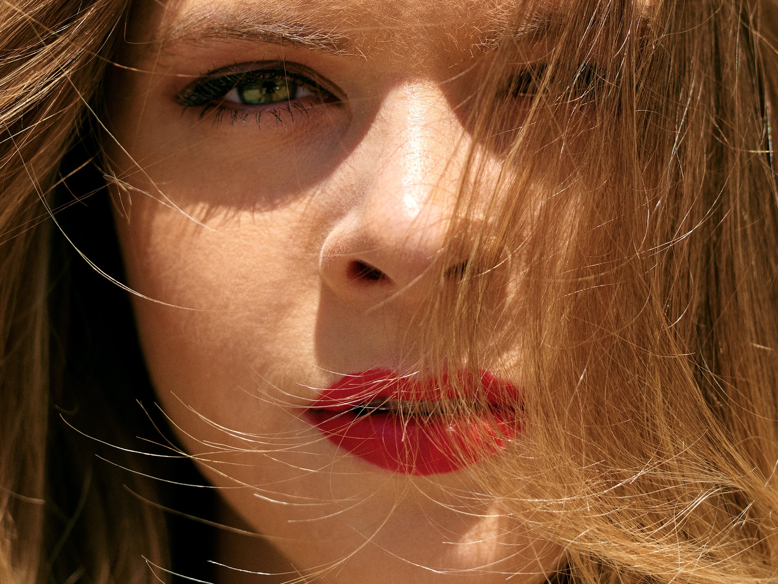 Close-up of a woman with long hair and one eye closed, wearing makeup, with sunlight hitting her face.