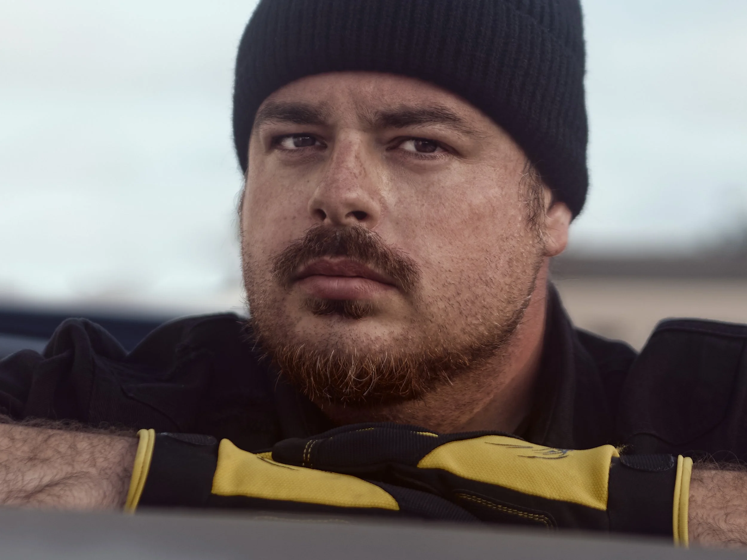 A close-up of a man wearing a black beanie and a black and yellow sports jersey, looking into the camera with a serious expression, outdoors with an overcast sky in the background.