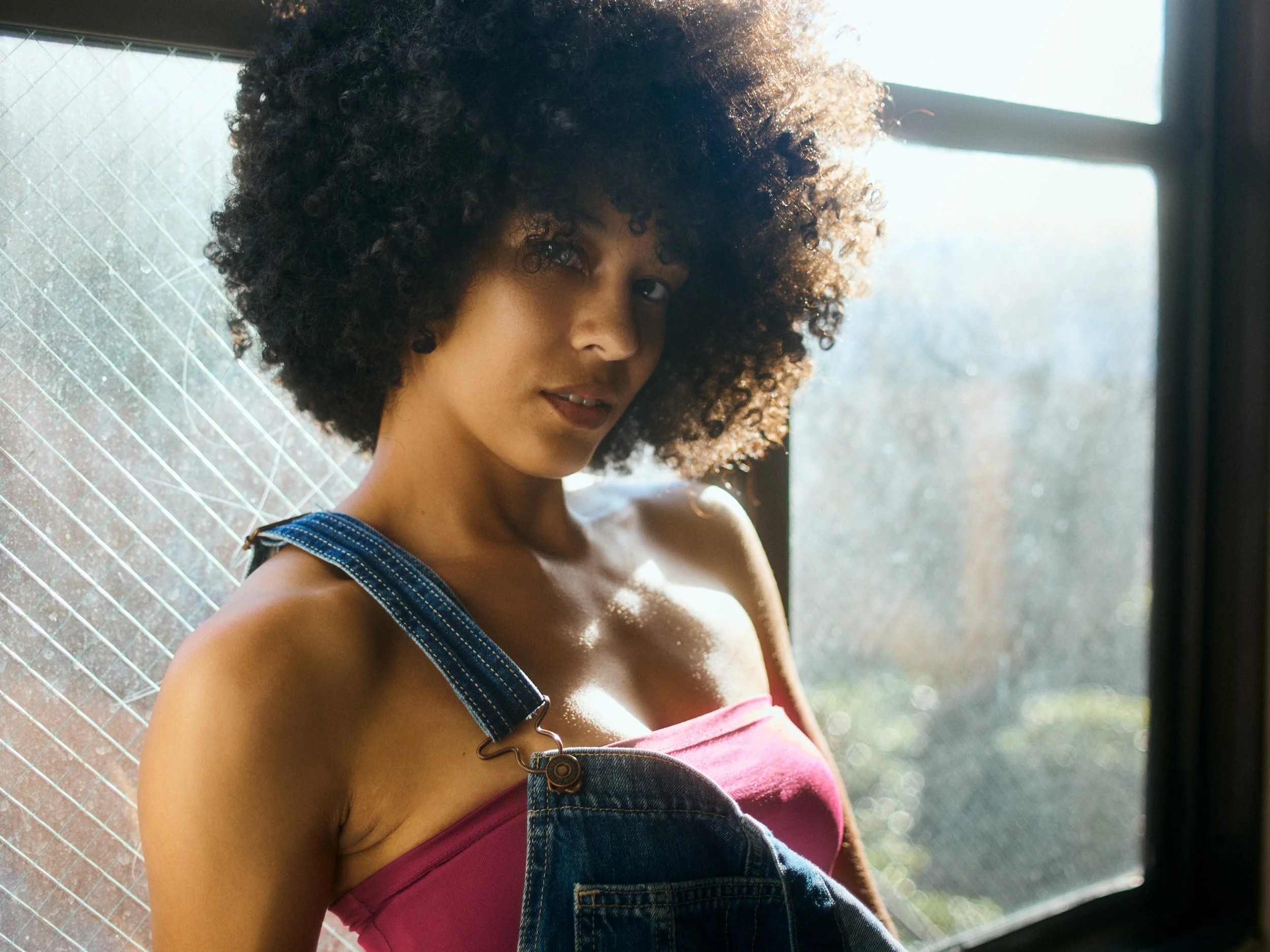 A woman with curly hair wearing a pink top and denim overalls, standing in front of a window with sunlight shining through, partially obscured by a patterned window covering.