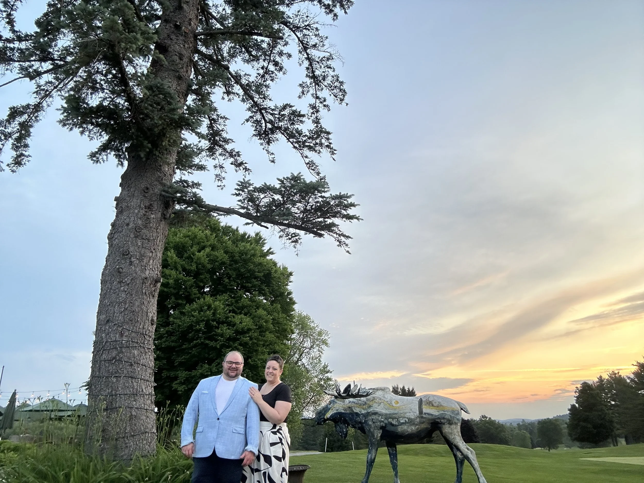 Two smiling people, a man in a light blue suit and a person in black, standing outside near a large tree at sunset. There is a decorative sculpture of a moose in the background.