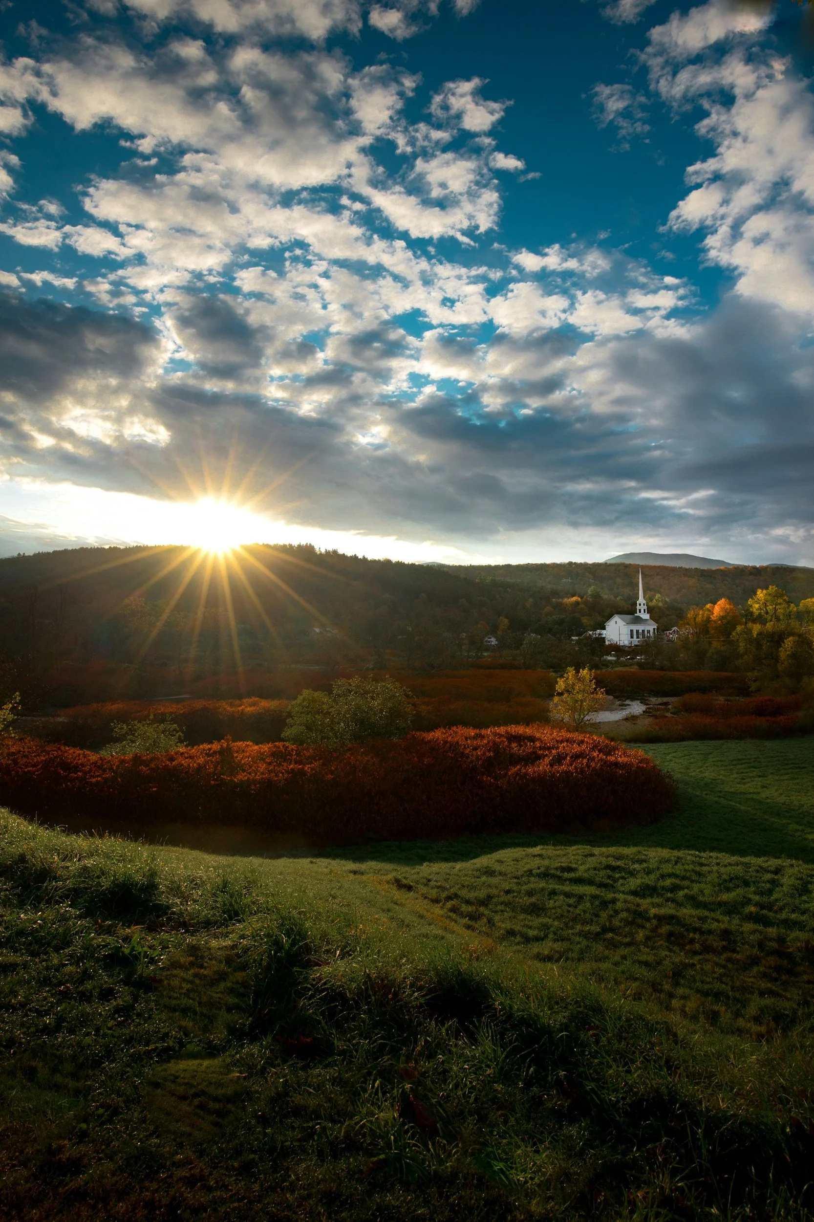 Sunset over a rural landscape with a white church on a hill and colorful autumn foliage in the foreground, under a partly cloudy sky.