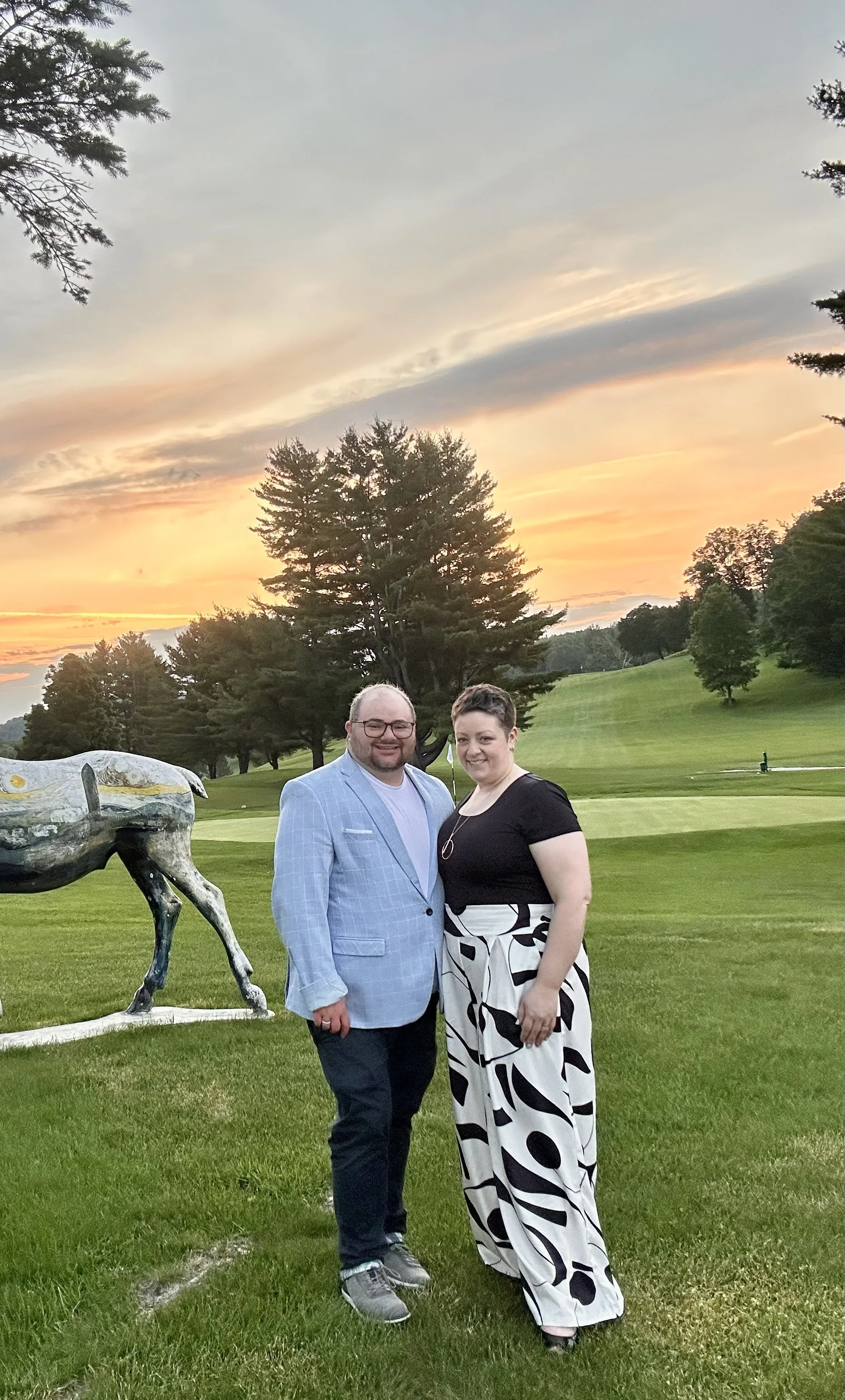 A man wearing a light blue checkered blazer, dark pants, and sneakers, and a person in a black top with a white and black patterned skirt, standing together on a grassy area at sunset with trees and a sculpture of a horse in the background.