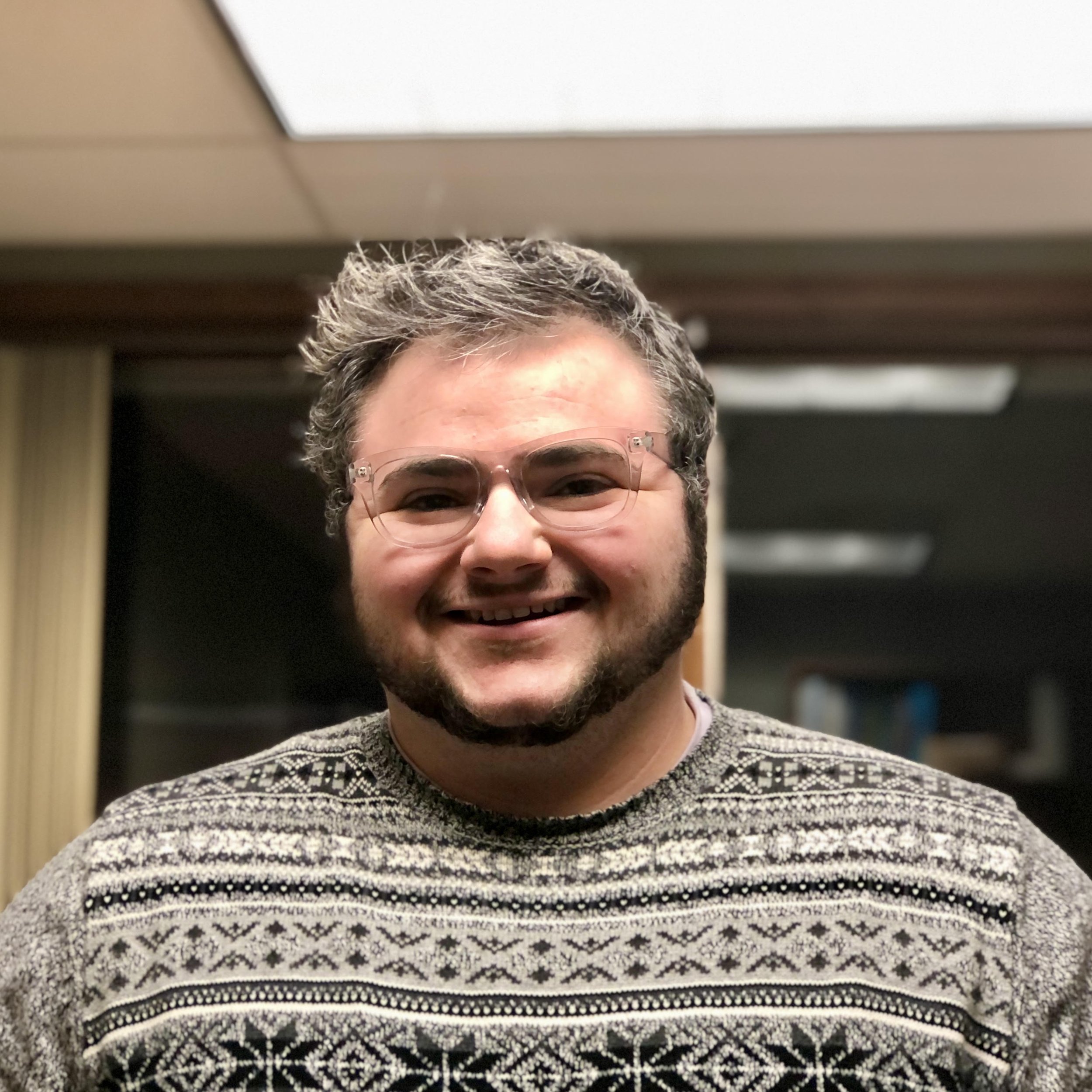 A person with gray and black hair smiling, wearing glasses and a patterned black and white sweater, photo taken indoors.