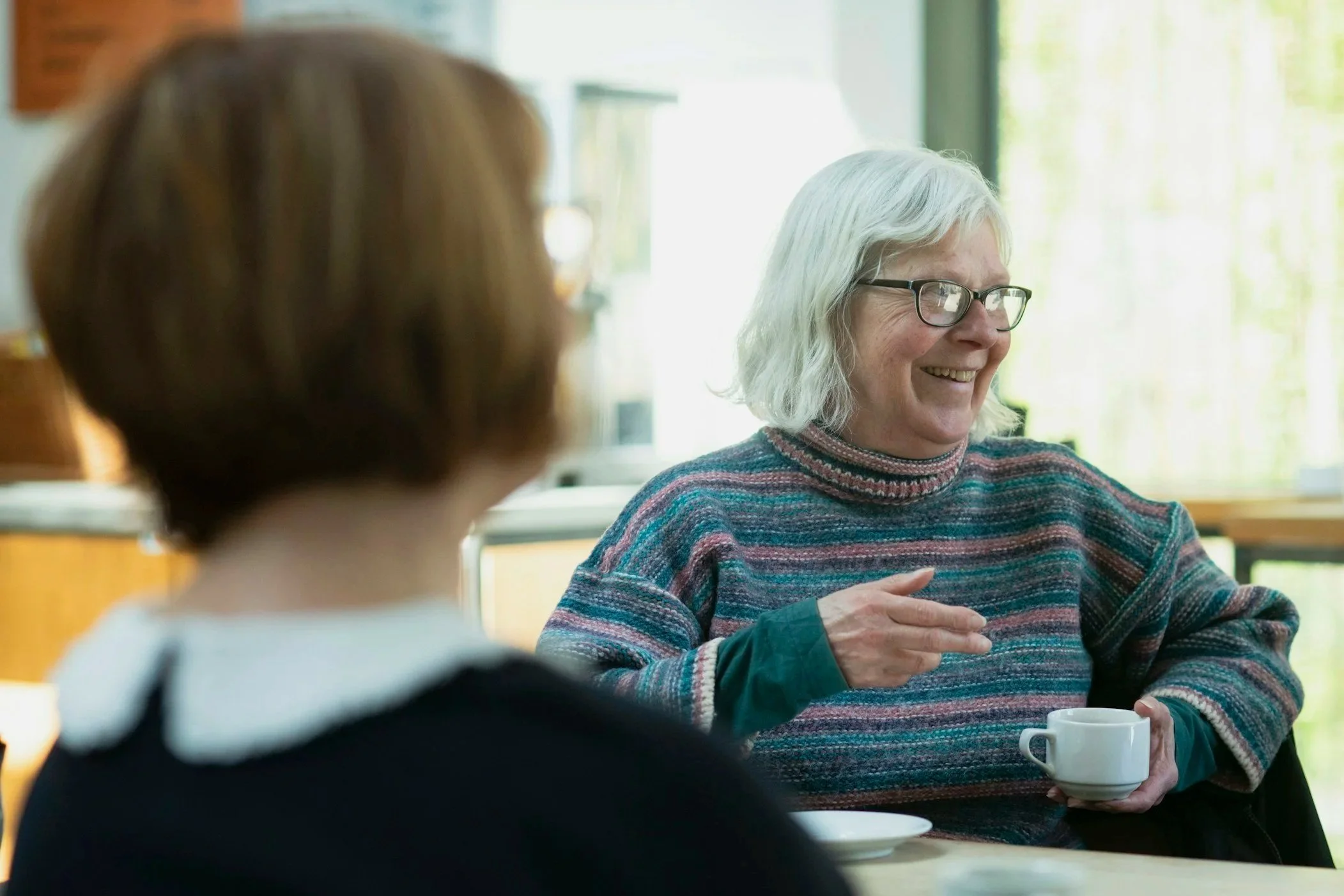 An elderly woman with gray hair and glasses smiling, holding a cup of coffee, sitting at a table in a bright room with large windows, with a younger person with brown hair in the foreground.