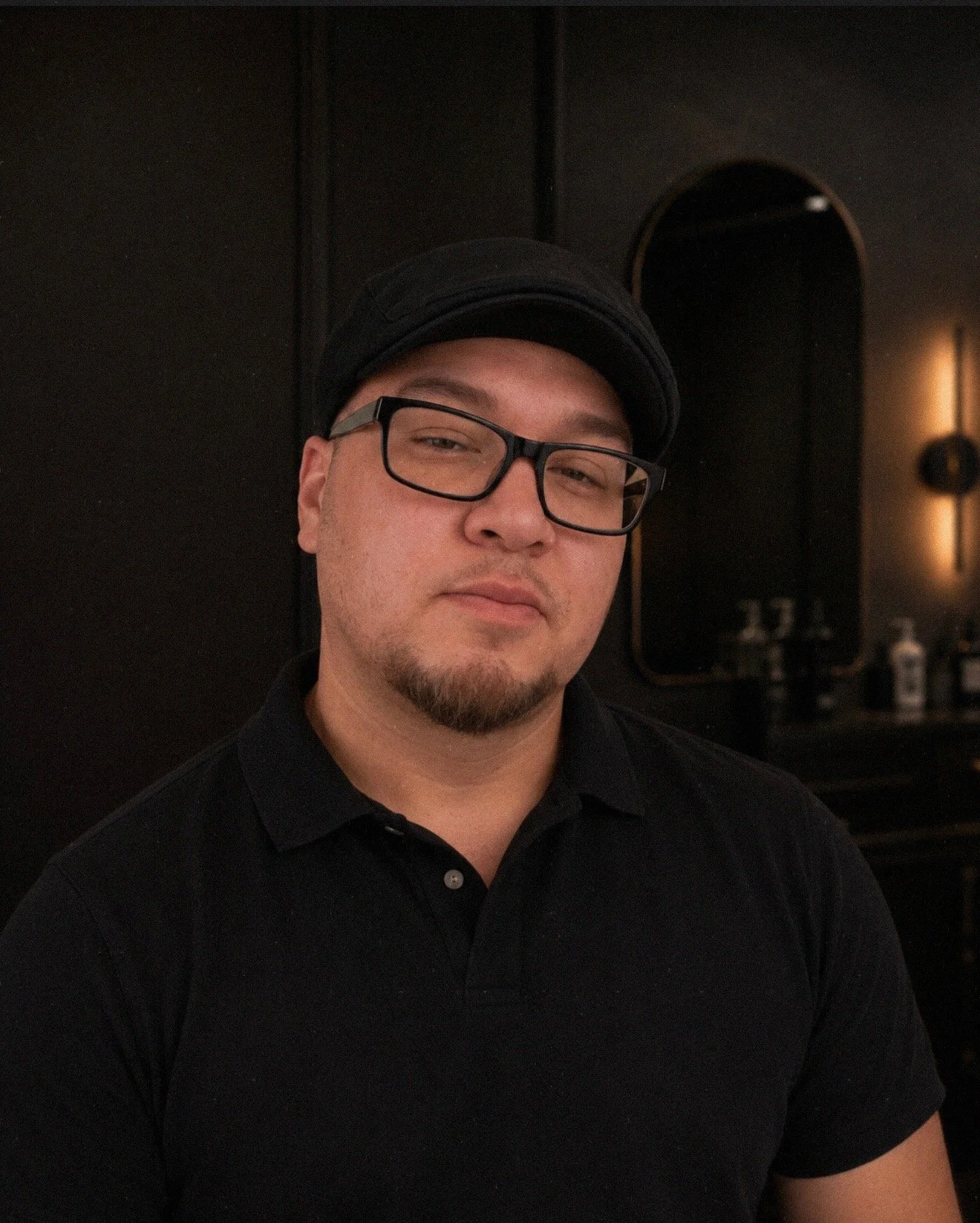 A man wearing glasses, a black cap, and a black polo shirt, posing indoors against a dark background with a mirror and bottles visible on a counter.