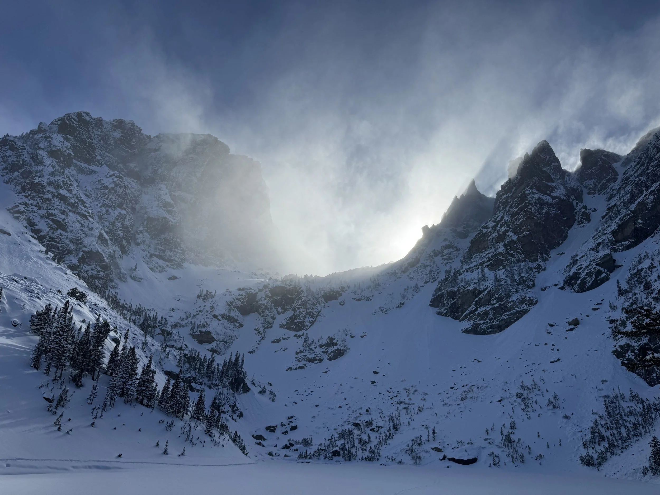 Snow-covered mountain range with rocky peaks and a few trees in the foreground, the sun shining behind the mountains with a partly cloudy sky.