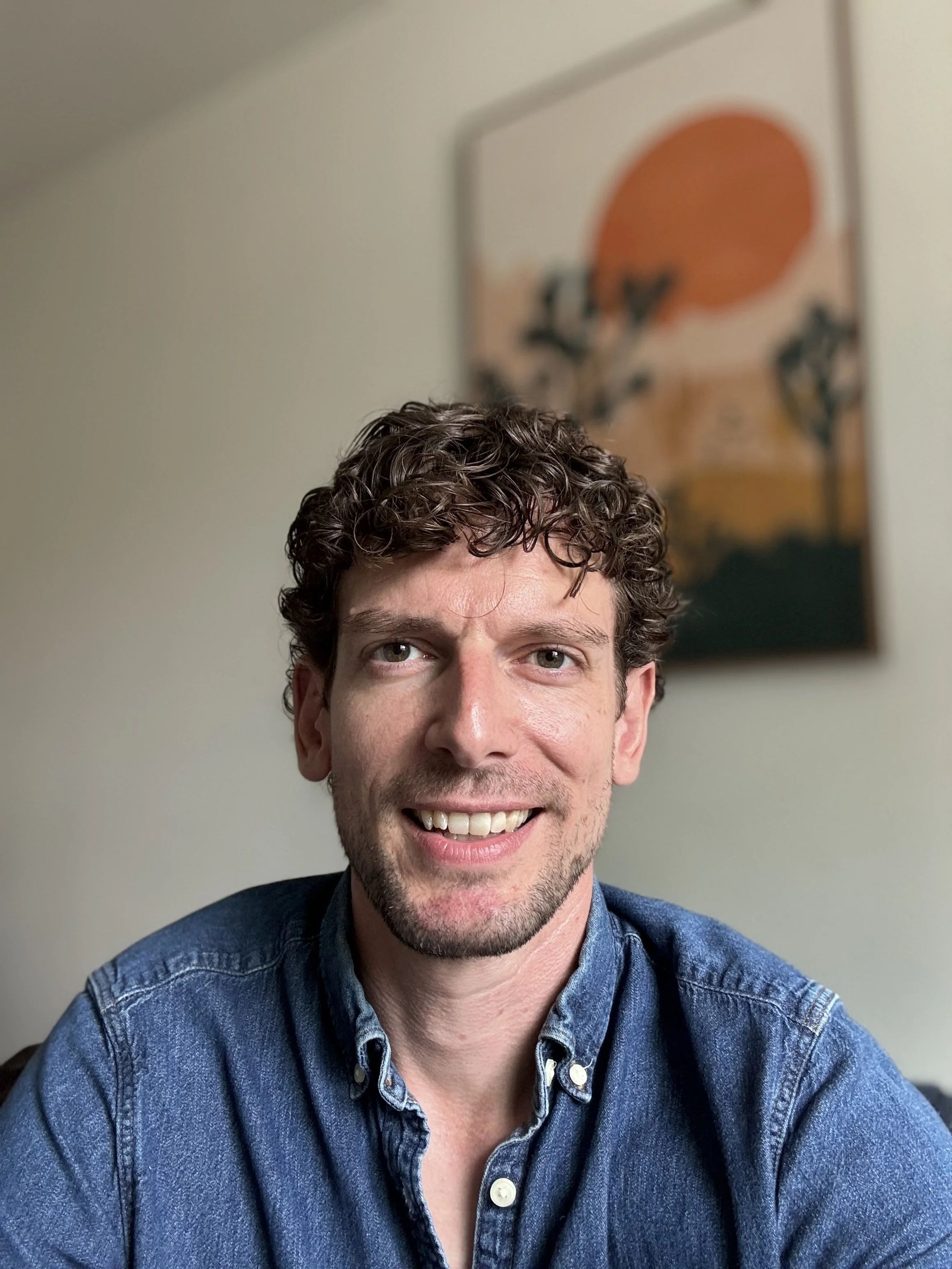 A man with curly brown hair and light skin smiling, wearing a denim shirt, sitting in front of a beige wall with abstract artwork in the background.