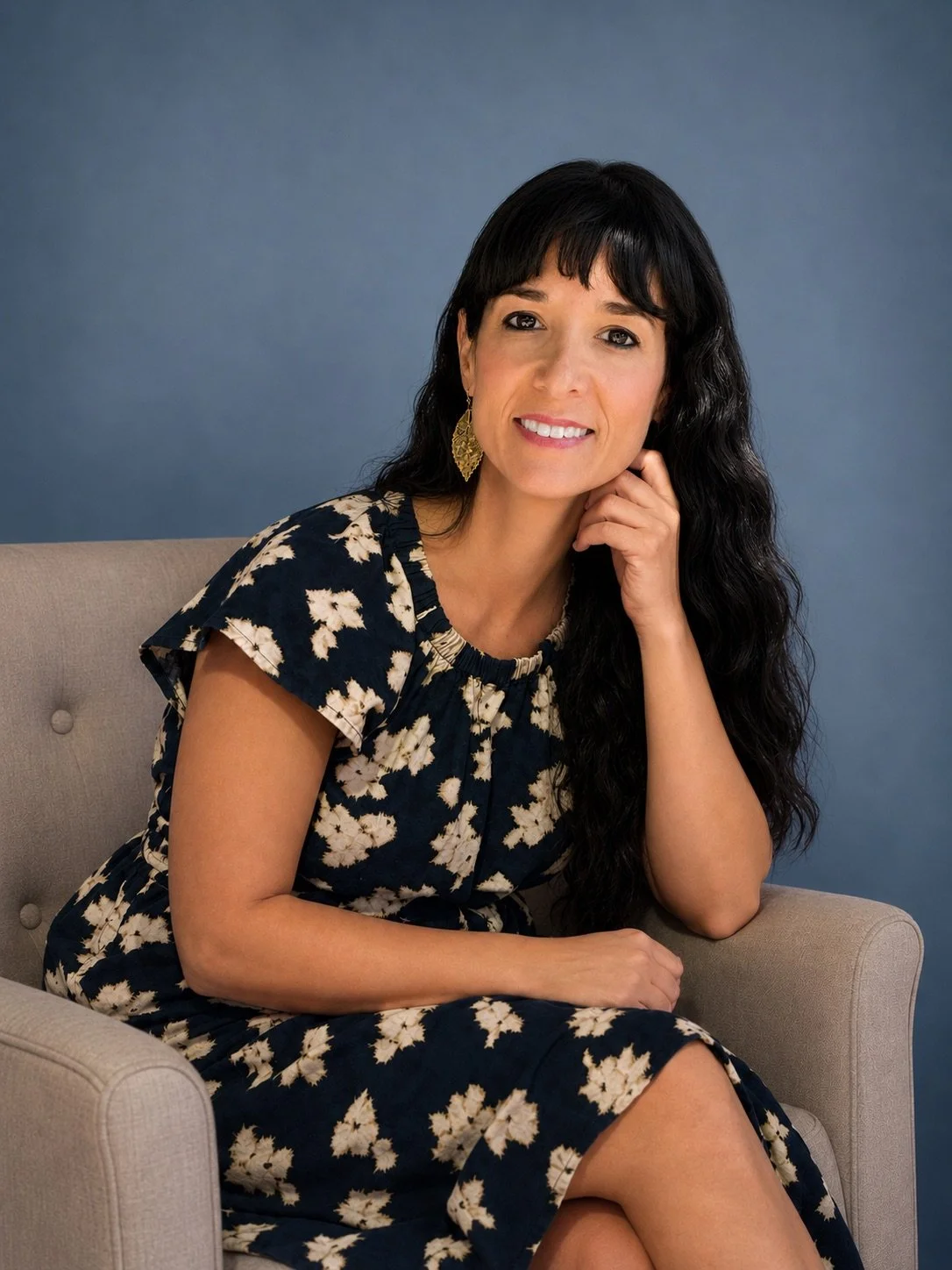 A woman with long black wavy hair, wearing a navy dress with white floral pattern, sitting on a beige armchair against a blue background, smiling and resting her chin on her right hand.