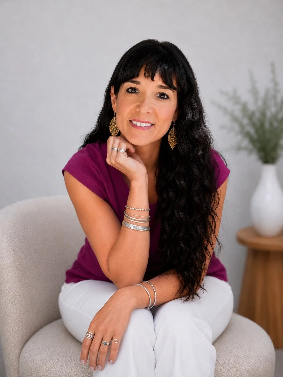 A woman with long, dark wavy hair wearing a magenta top and white pants, sitting on a beige chair in a neutral room with a grey wall and a white vase with greenery in the background.