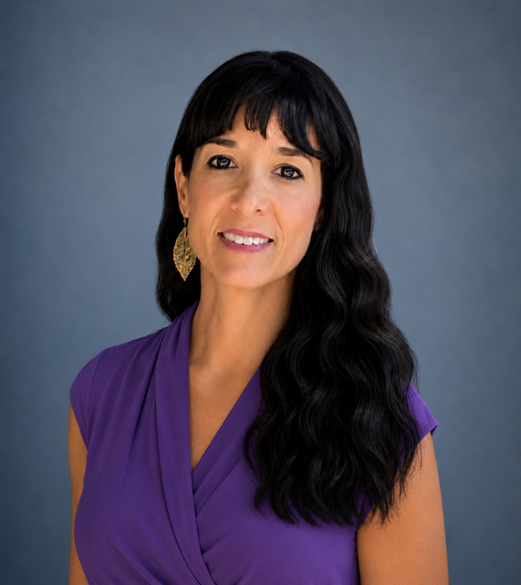 A woman with long black wavy hair, wearing a purple top and gold earrings, smiling against a blue background.