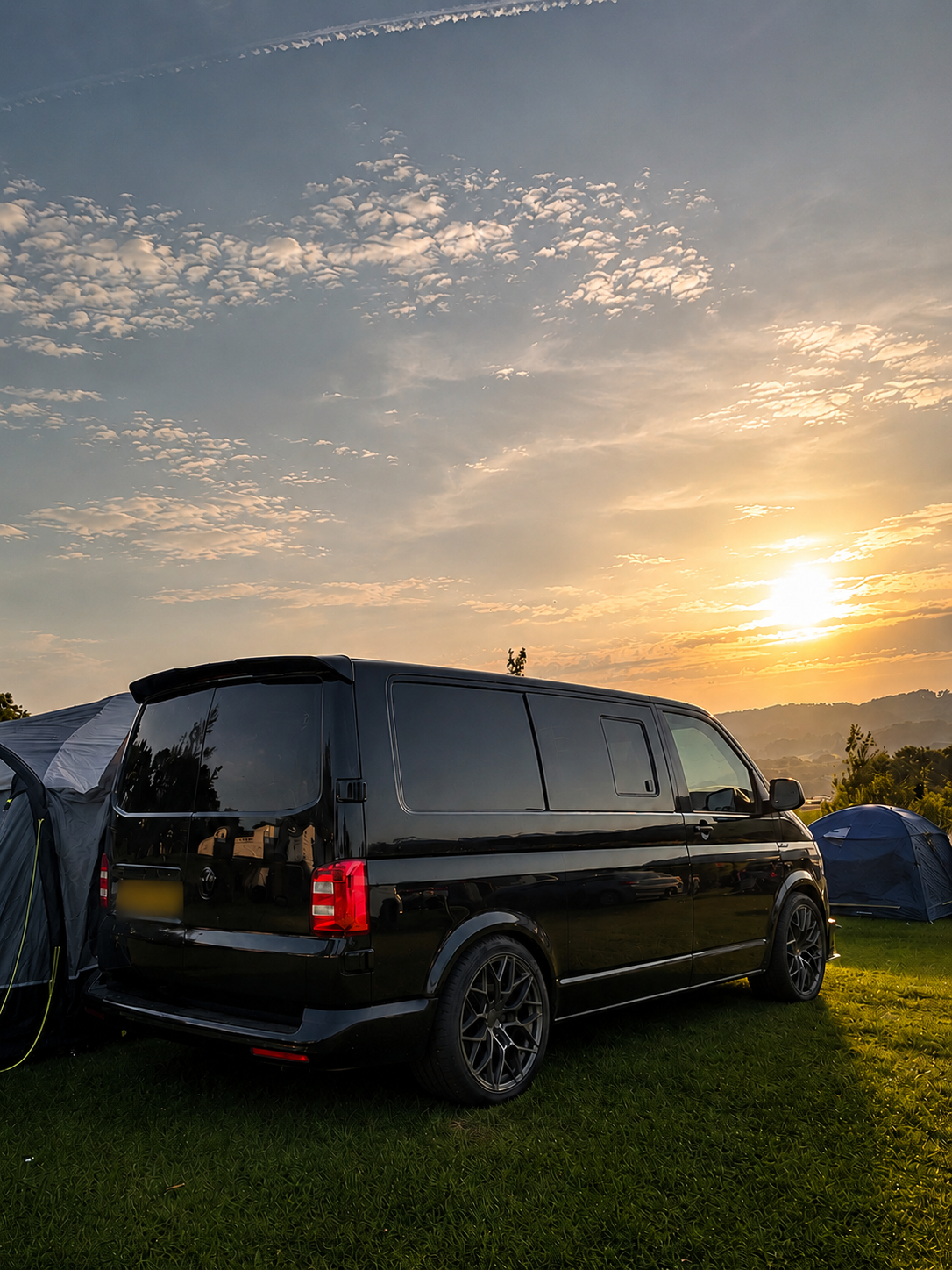 A black van parked on a grassy field during sunset with tents nearby and hills in the background.