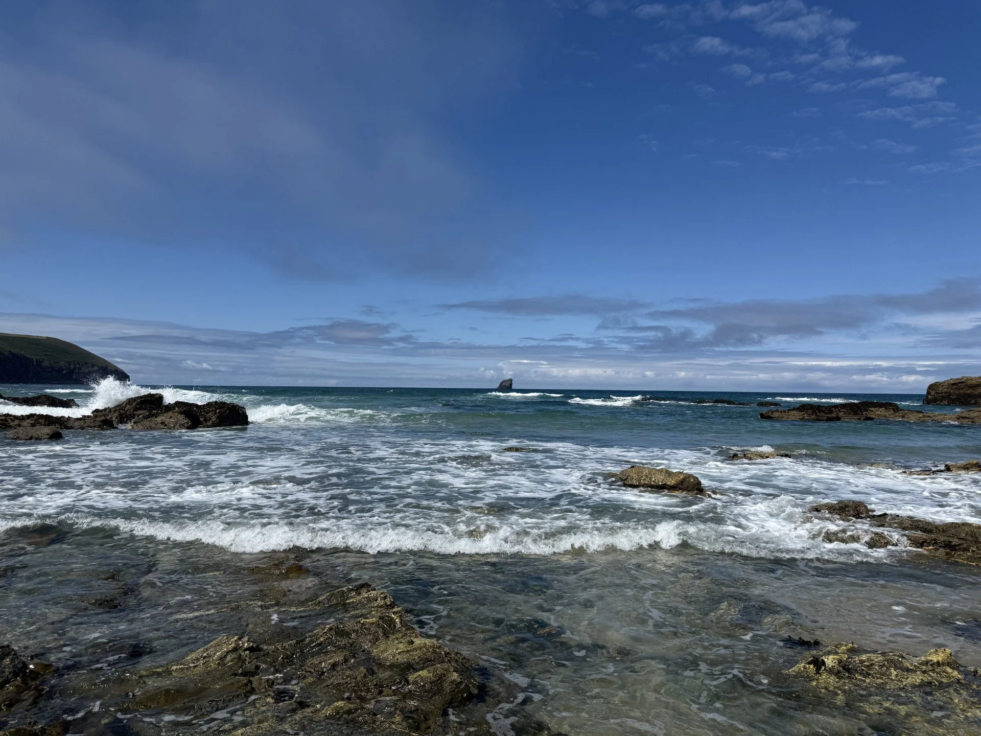 A view of a rocky coastline with the ocean crashing against the rocks under a blue sky with clouds.