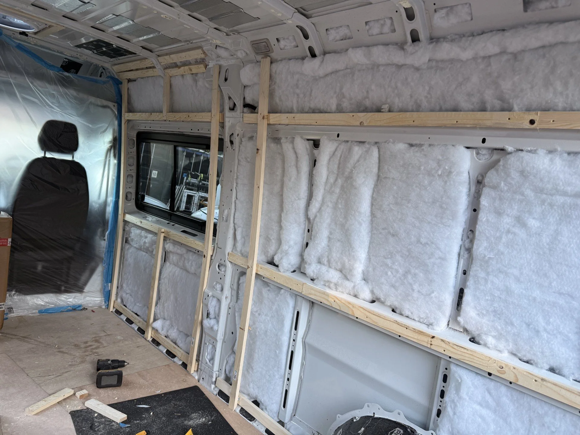 Interior of a van with insulation and wooden framing installed, with tools and materials on the floor.