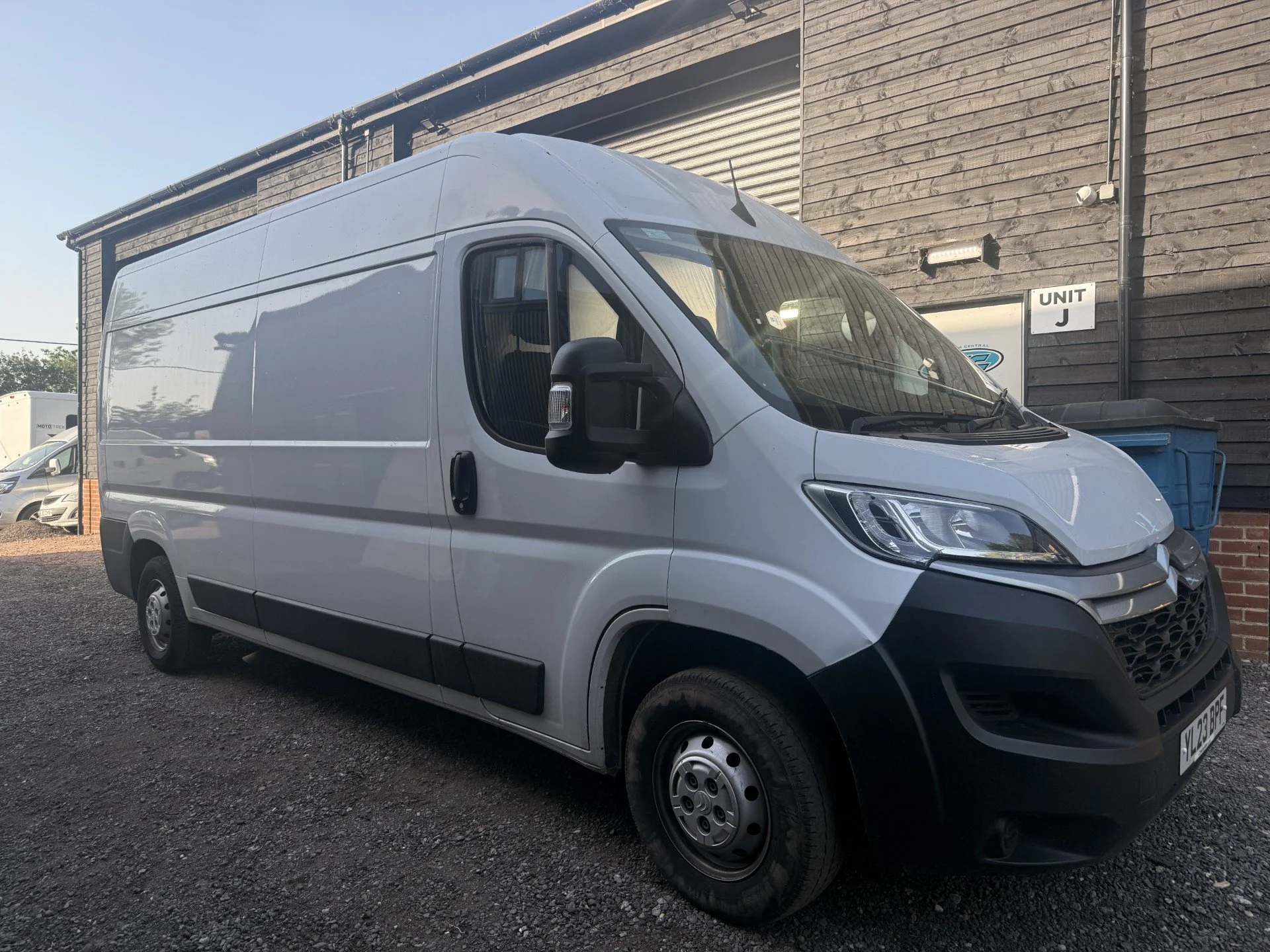 A white commercial van parked outside a industrial building with wooden walls. The van's front end and part of the driver's side are visible, with a focus on the passenger side and the building in the background.
