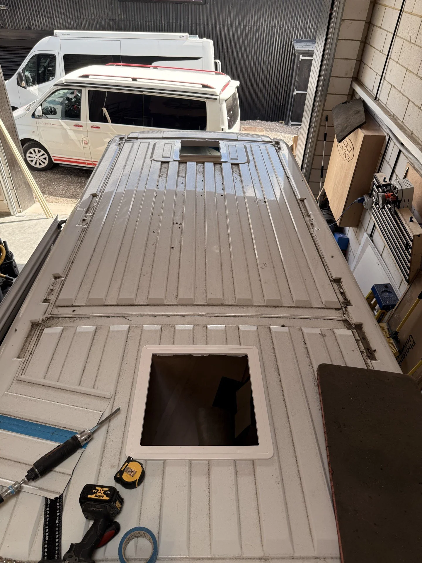 Inside a workshop with tools, a cordless drill, tape measure, and a level on a white work surface. The background shows the foot of a van or trailer with an open hatch and a small window.