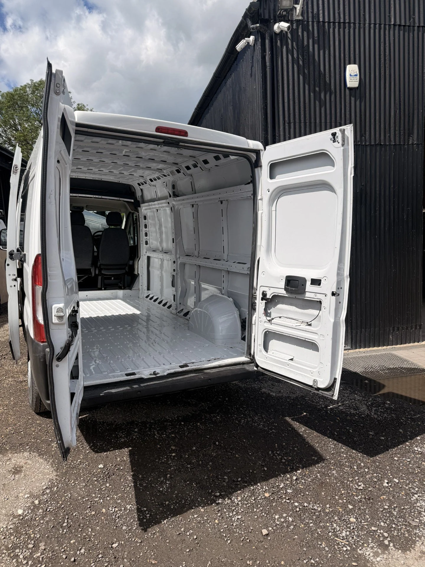 Rear view of an empty white cargo van with open doors, parked on gravel, with a black building and cloudy sky in the background.